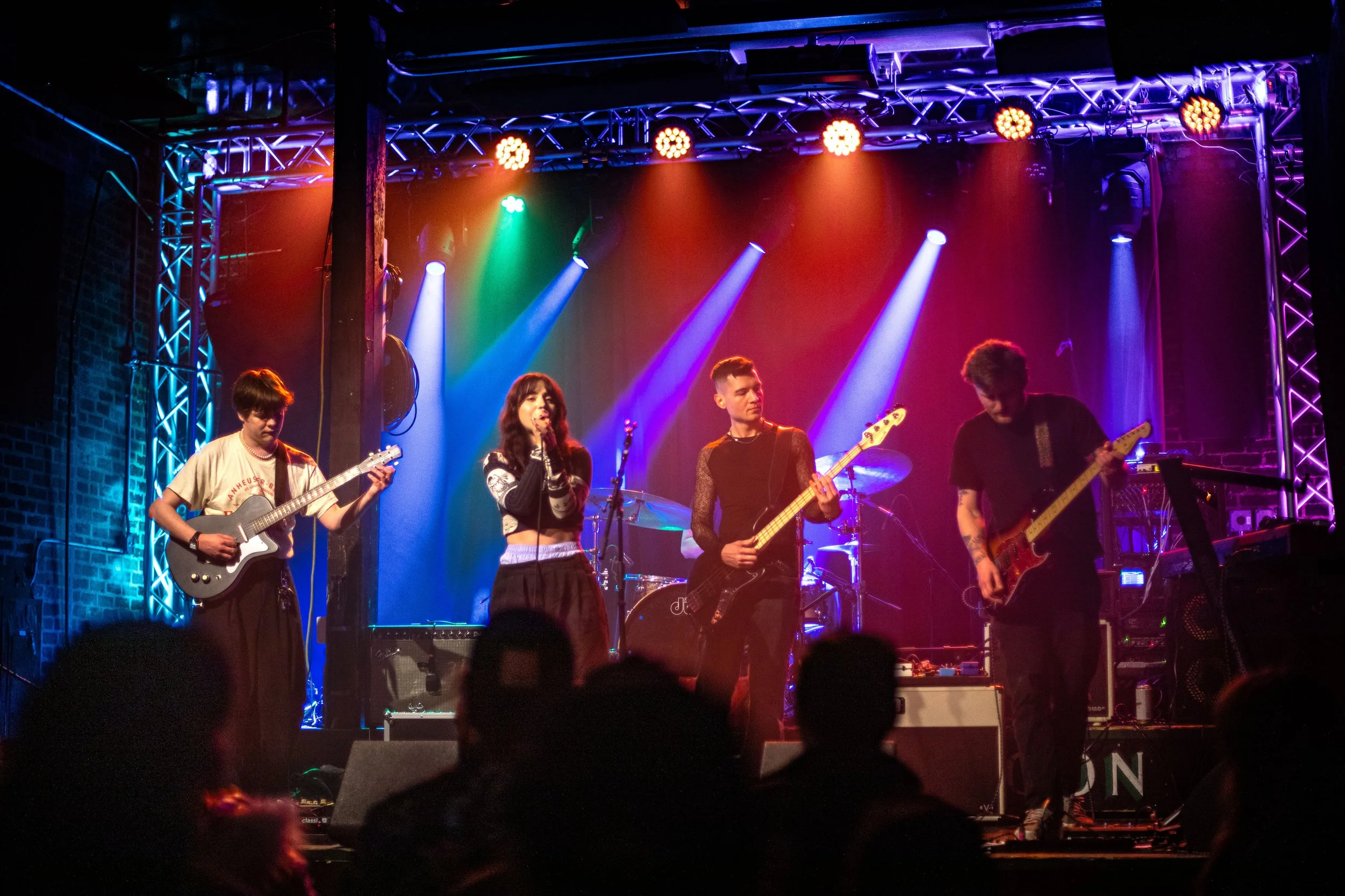 A band of five musicians performing on stage with colorful lighting, including a female vocalist at the center and four instrumentalists playing guitar and drums.