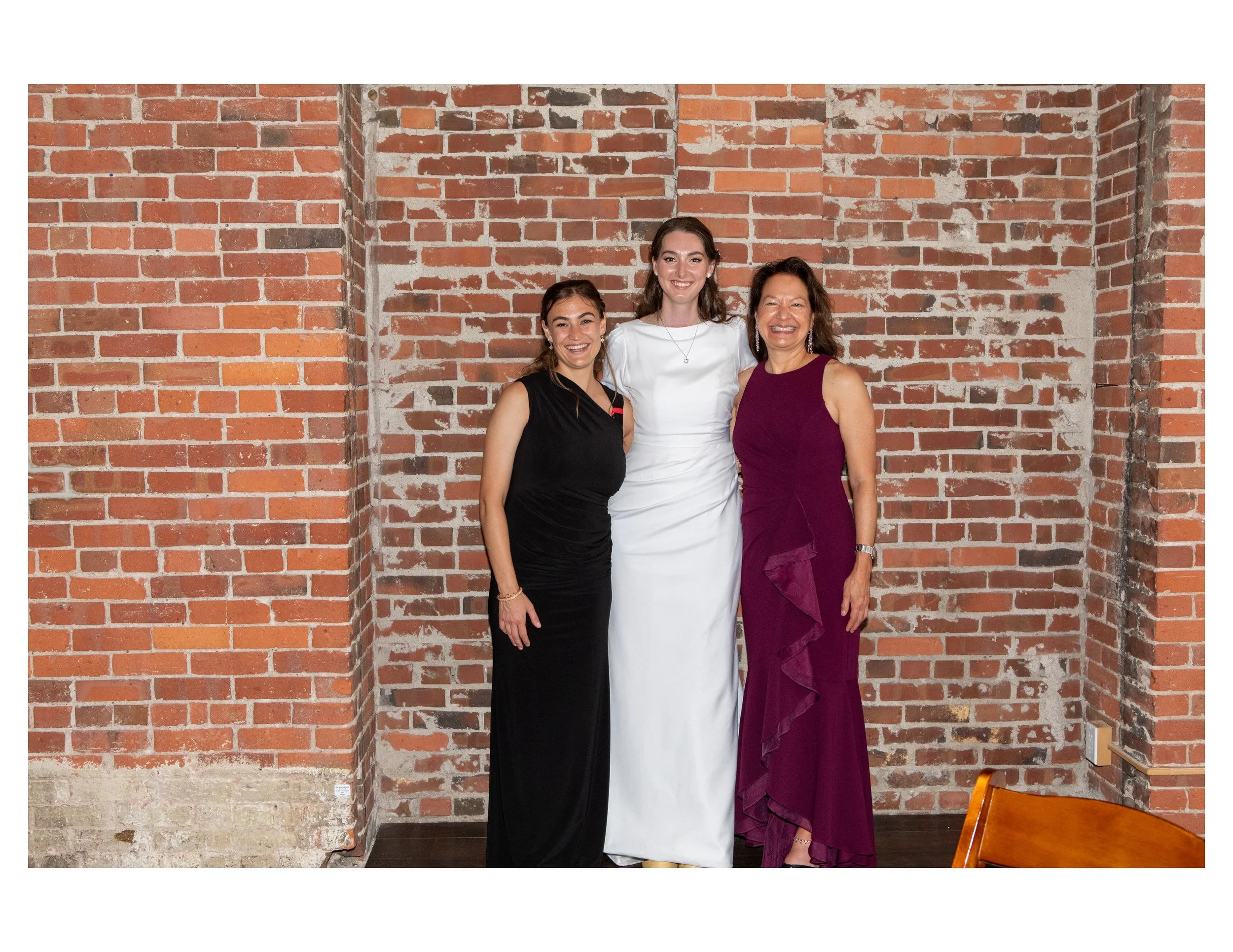 Three women smiling and posing against a red brick wall. The woman on the left is wearing a black dress, the woman in the middle is wearing a white dress, and the woman on the right is wearing a purple dress.