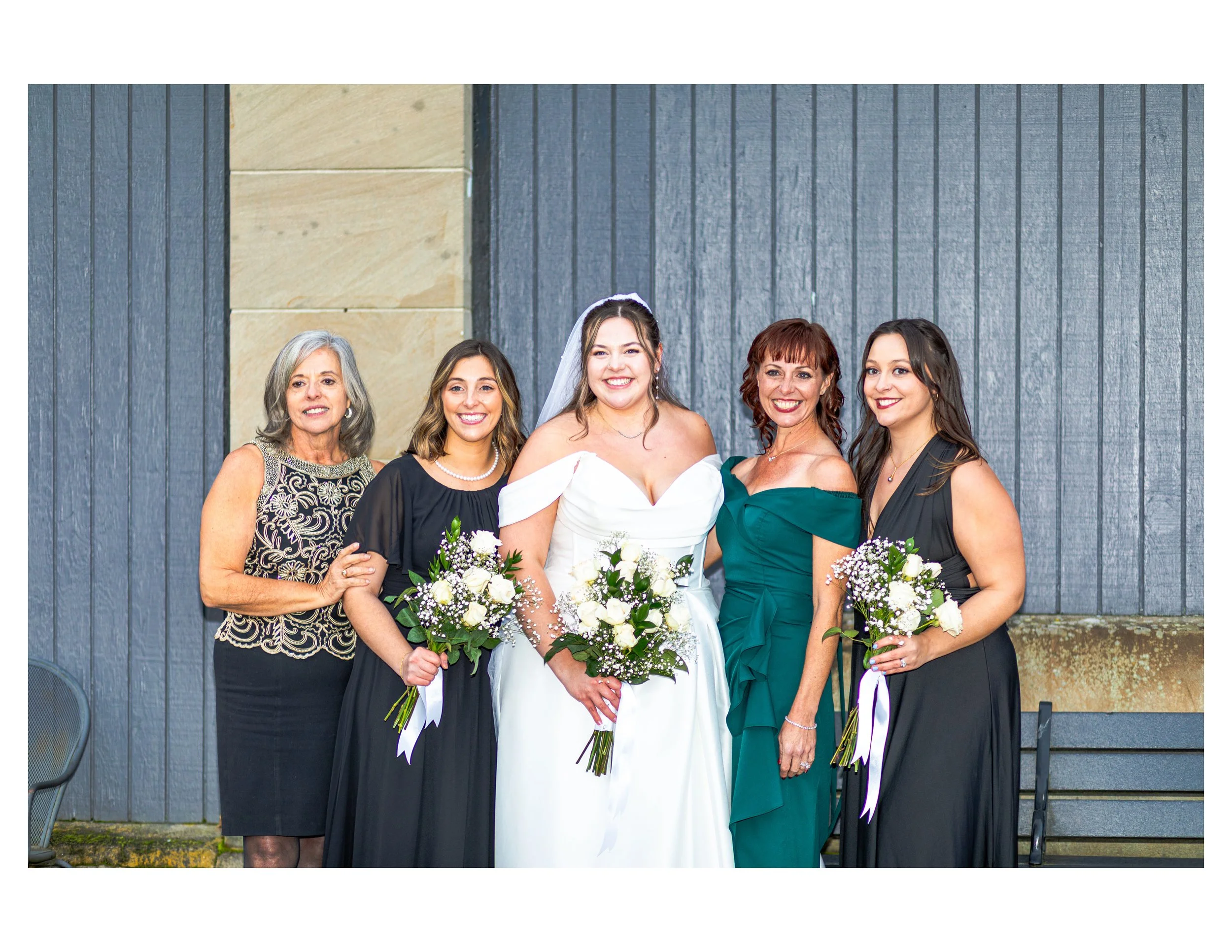 Group of five women, including a bride in a white dress, standing in front of a blue wooden wall, holding bouquets of white flowers with greenery, smiling for the photo.