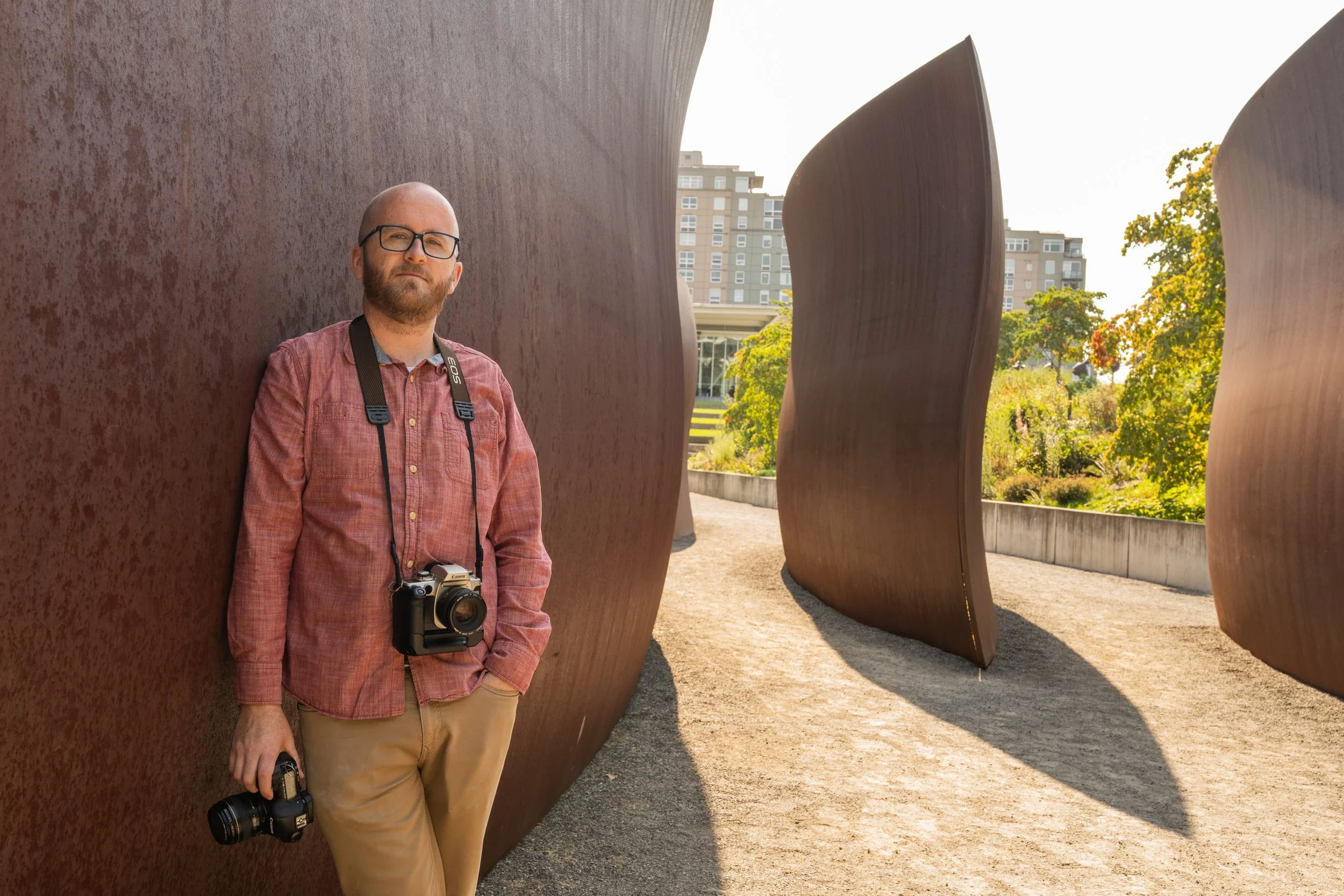 A man with glasses and a beard, wearing a pink button-up shirt with a camera hanging around his neck, standing outdoors beside rust-colored metal sculptures with a cityscape and trees in the background.