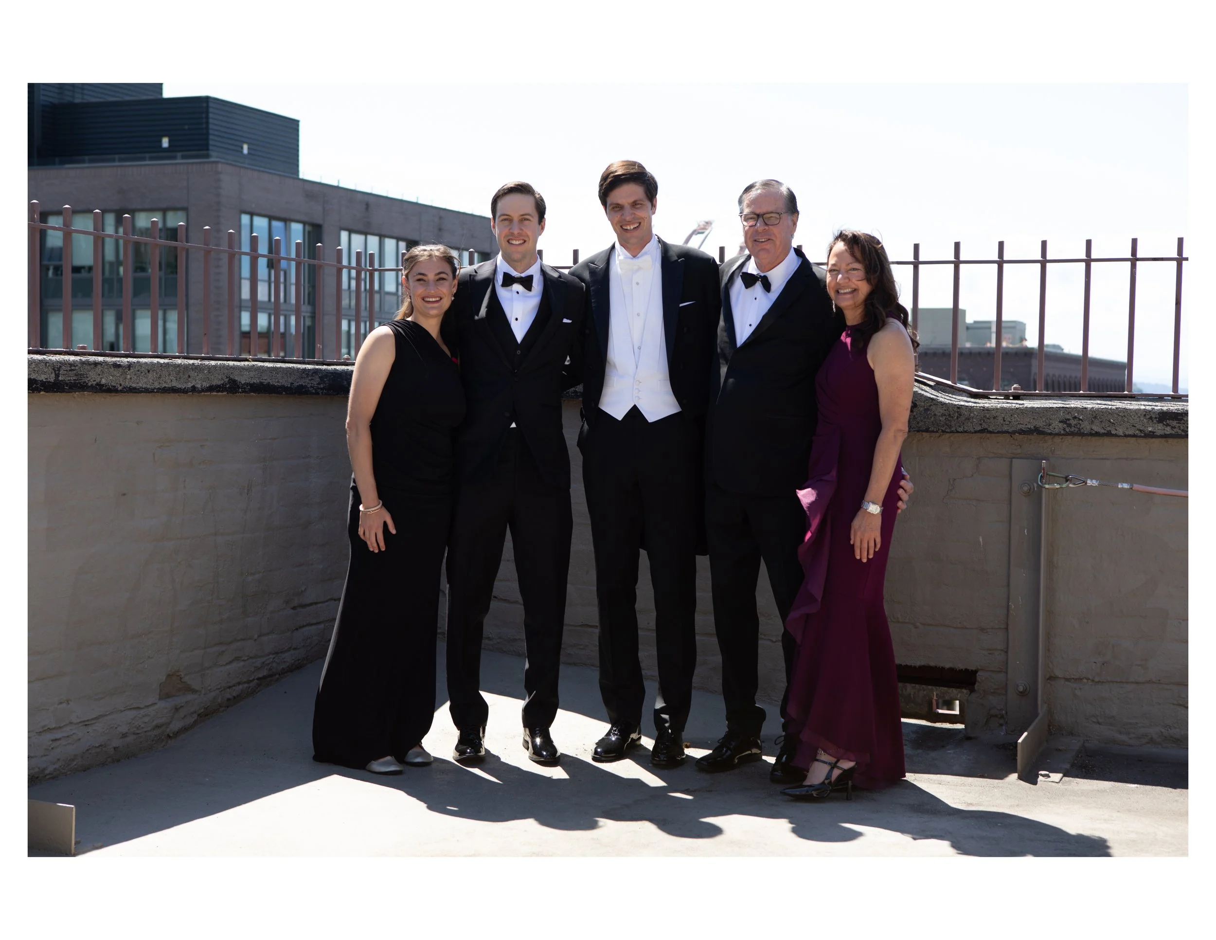 Five people dressed in formal attire standing on a rooftop balcony, smiling, with city buildings in the background.