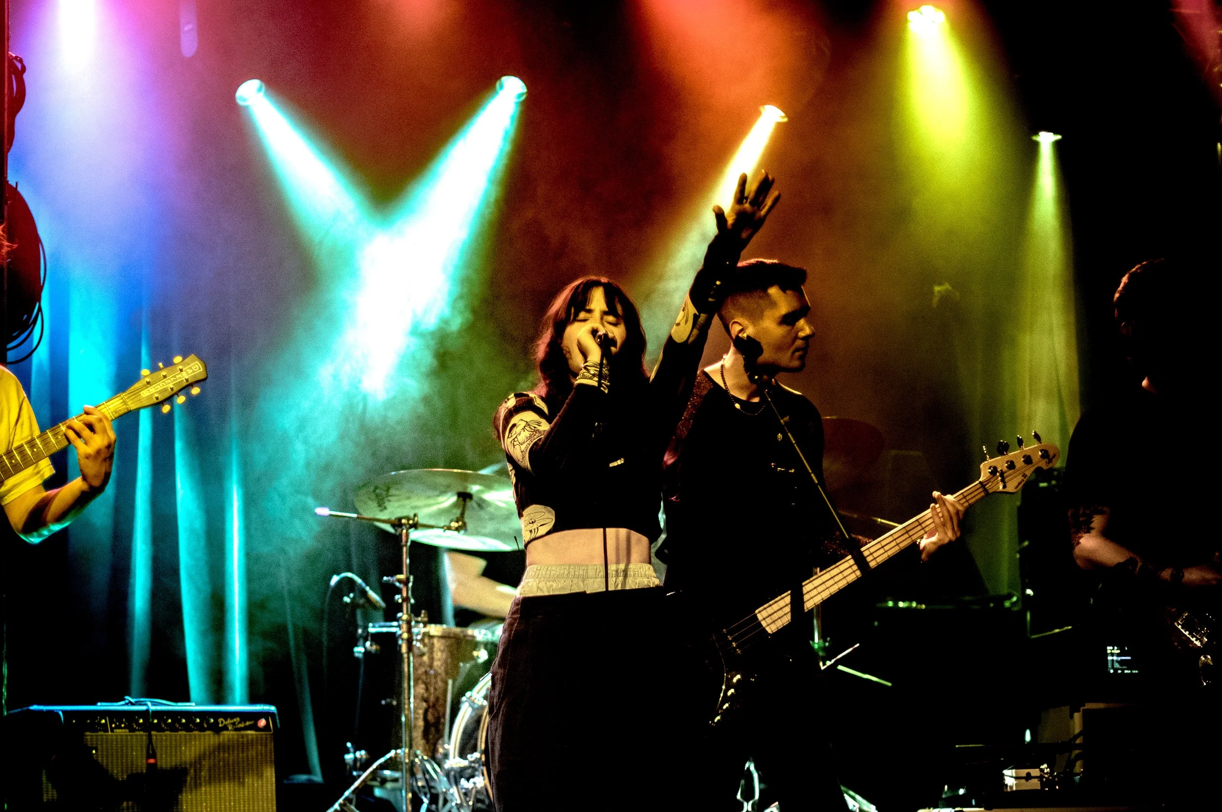 A band performing on stage with colorful lights, featuring a female lead singer with her eyes closed and her hand raised, a guitarist beside her, and a drummer in the background.