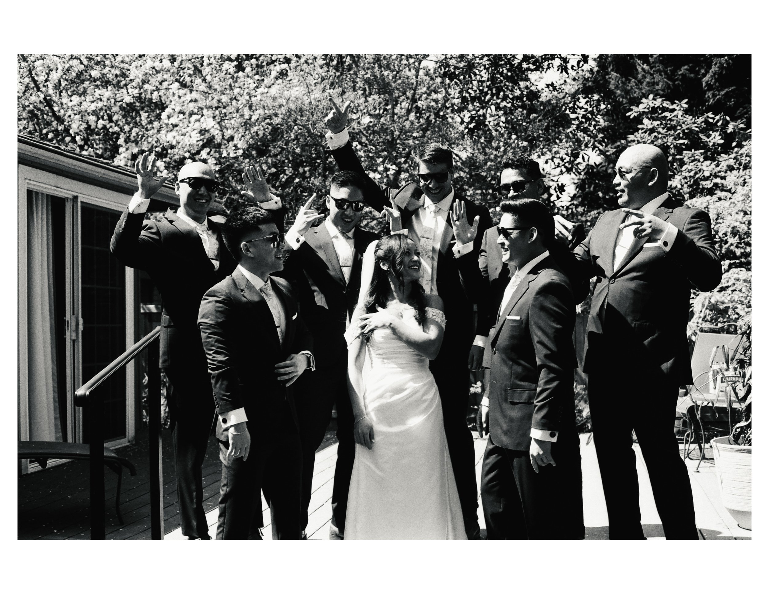 Group of people in suits at a wedding, some wearing sunglasses, smiling, and raising their hands, with a woman in a wedding dress in the center, outdoors with trees in the background.