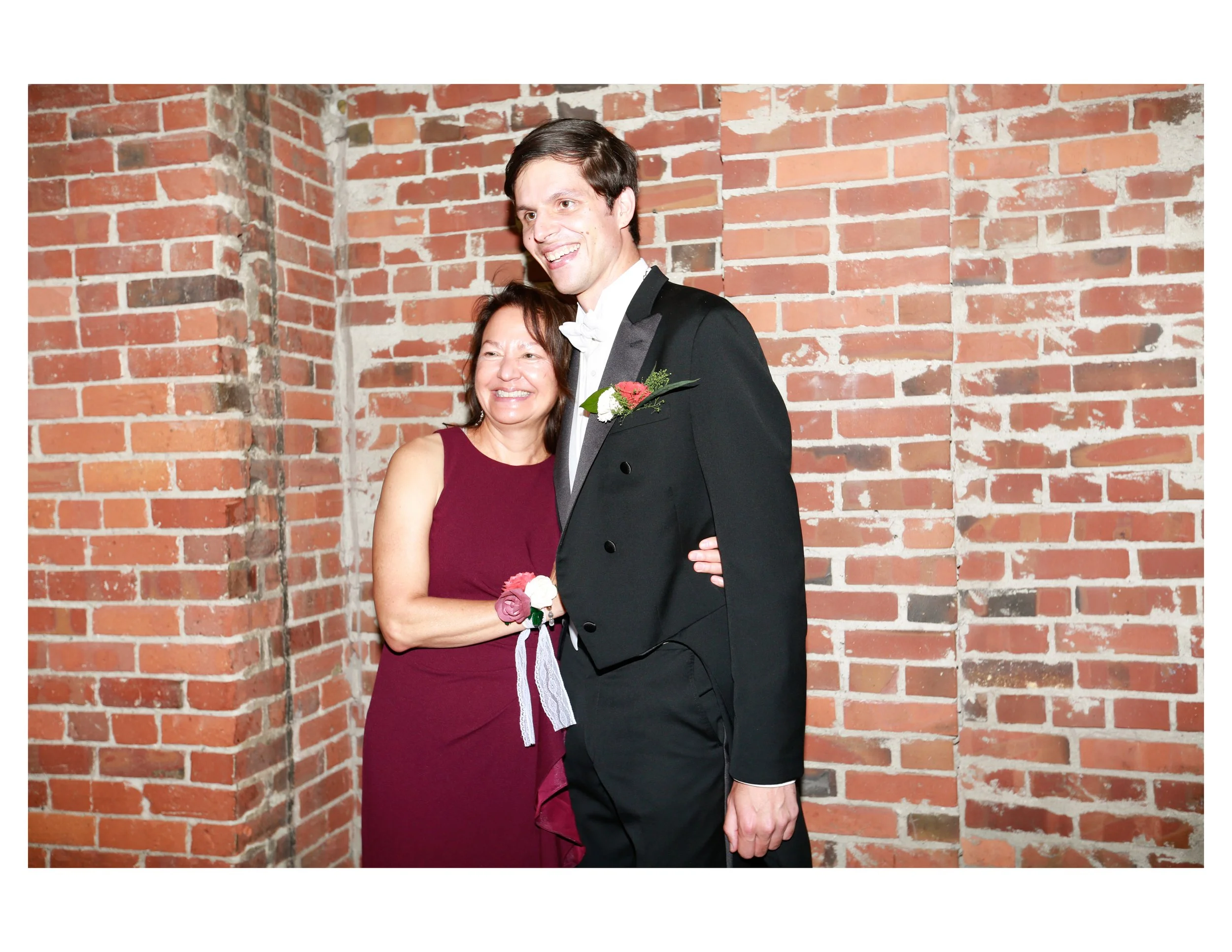 A man and woman smiling and embracing each other at a celebration, standing against a brick wall. The man is wearing a black tuxedo with a white shirt and bow tie, and the woman is in a sleeveless burgundy dress, holding a bouquet of flowers.