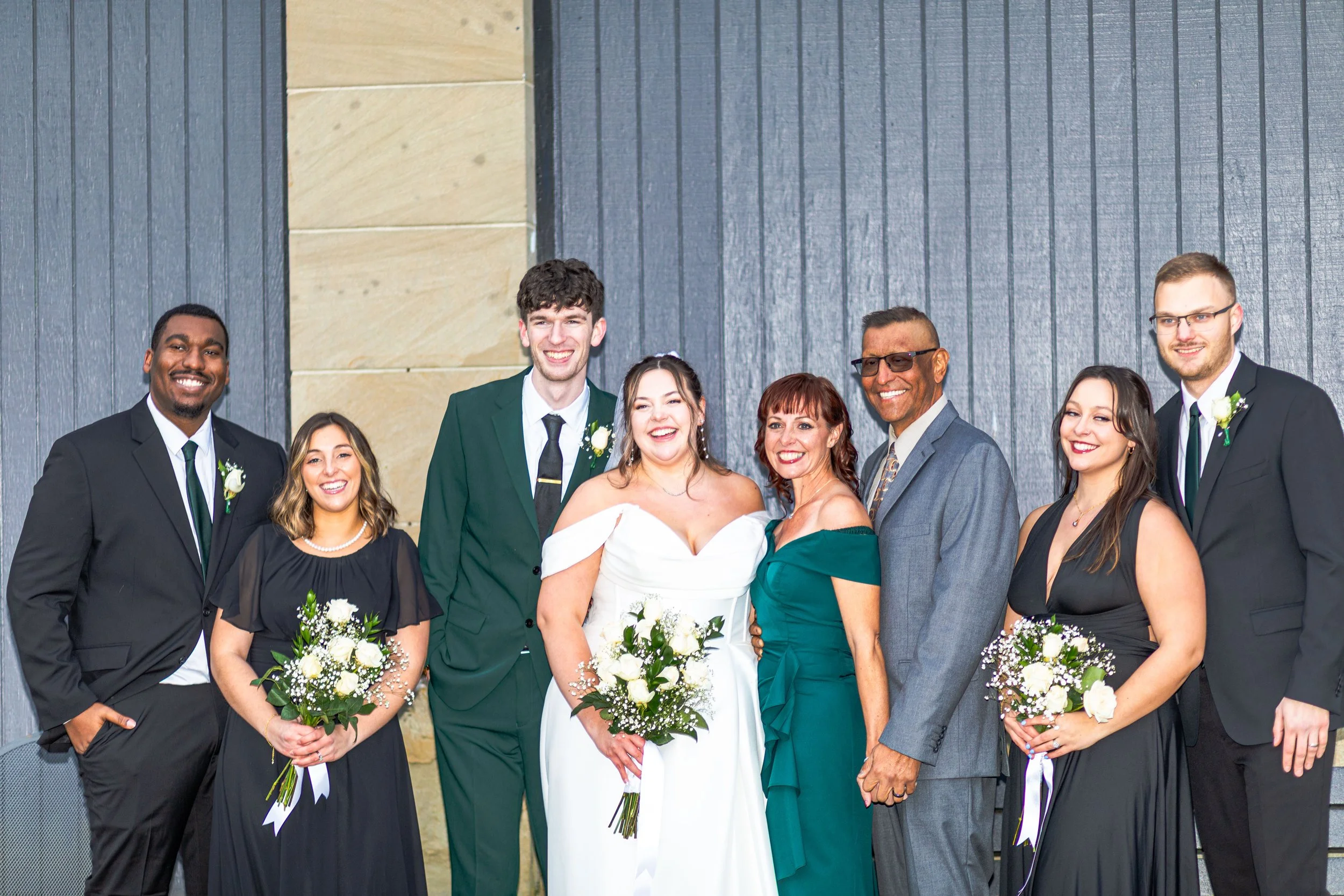 Group of people at a wedding celebration, including the bride in a white dress holding a bouquet, surrounded by family and friends dressed in formal attire, standing against a decorative wall.