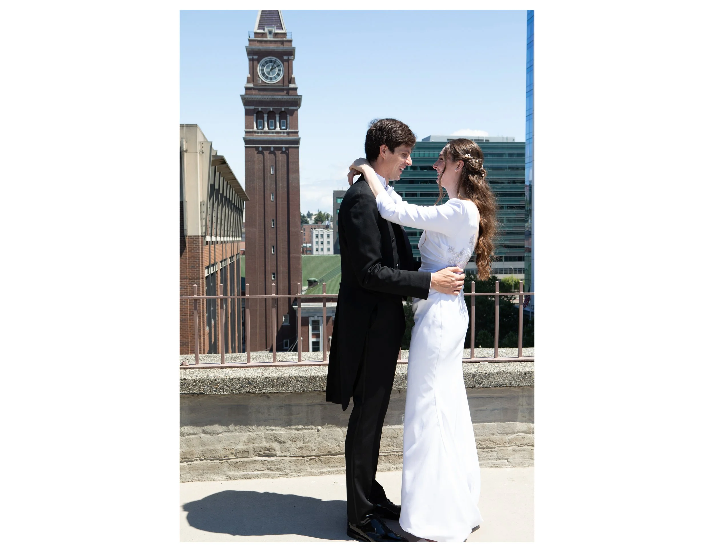 A man in a black suit and a woman in a white dress are standing close together, holding each other, and smiling at each other on a rooftop with a cityscape background, including a tall clock tower and modern buildings, under a clear blue sky.