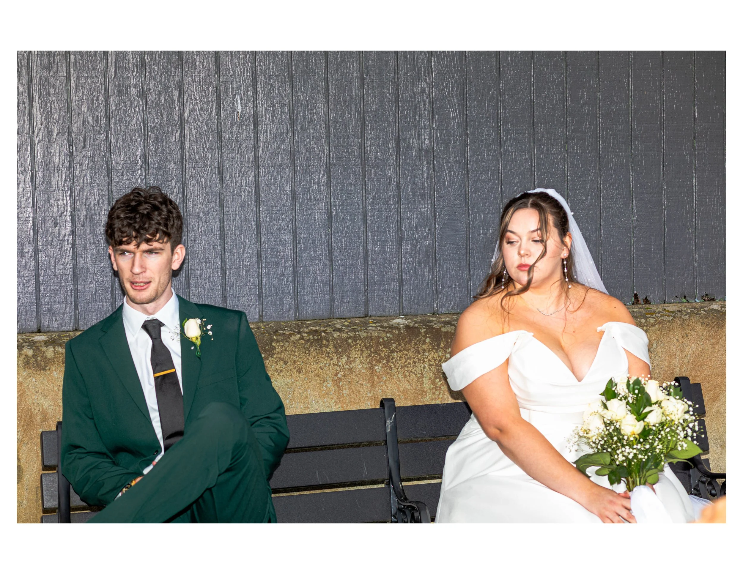 A young man in a green suit and a young woman in a white wedding dress sitting on a black bench in front of a dark gray wooden wall during a wedding ceremony, with the woman holding a bouquet of white and green flowers.