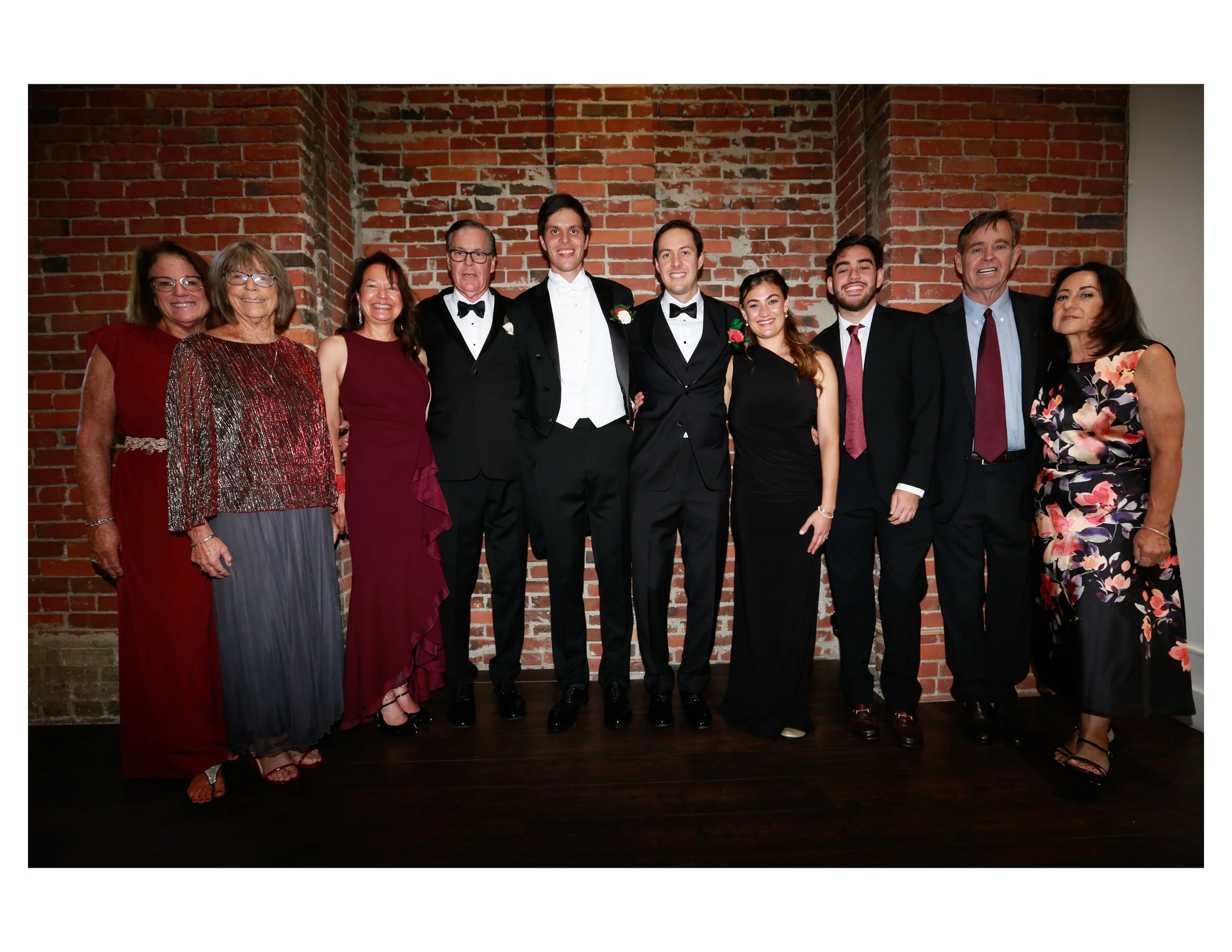 Group of ten people dressed in formal attire standing in front of a brick wall, smiling for a photo.