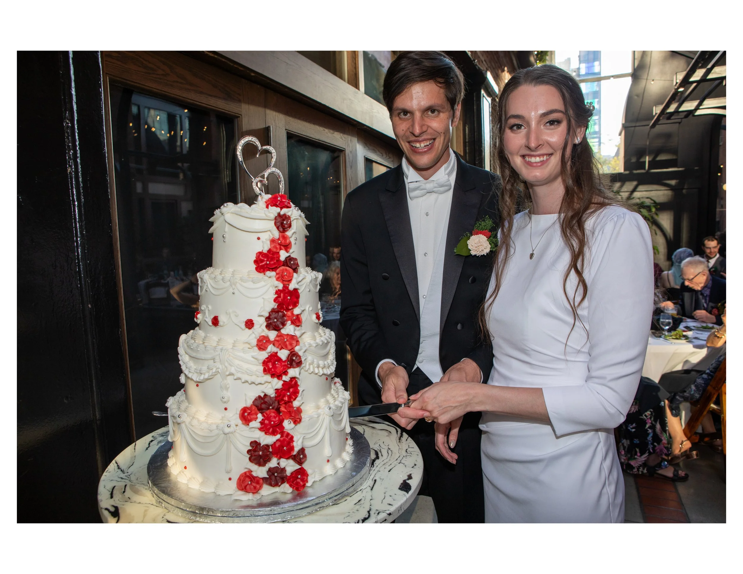 A smiling groom and bride at a wedding reception, standing beside a multi-tiered wedding cake decorated with red flowers and a heart-shaped cake topper.