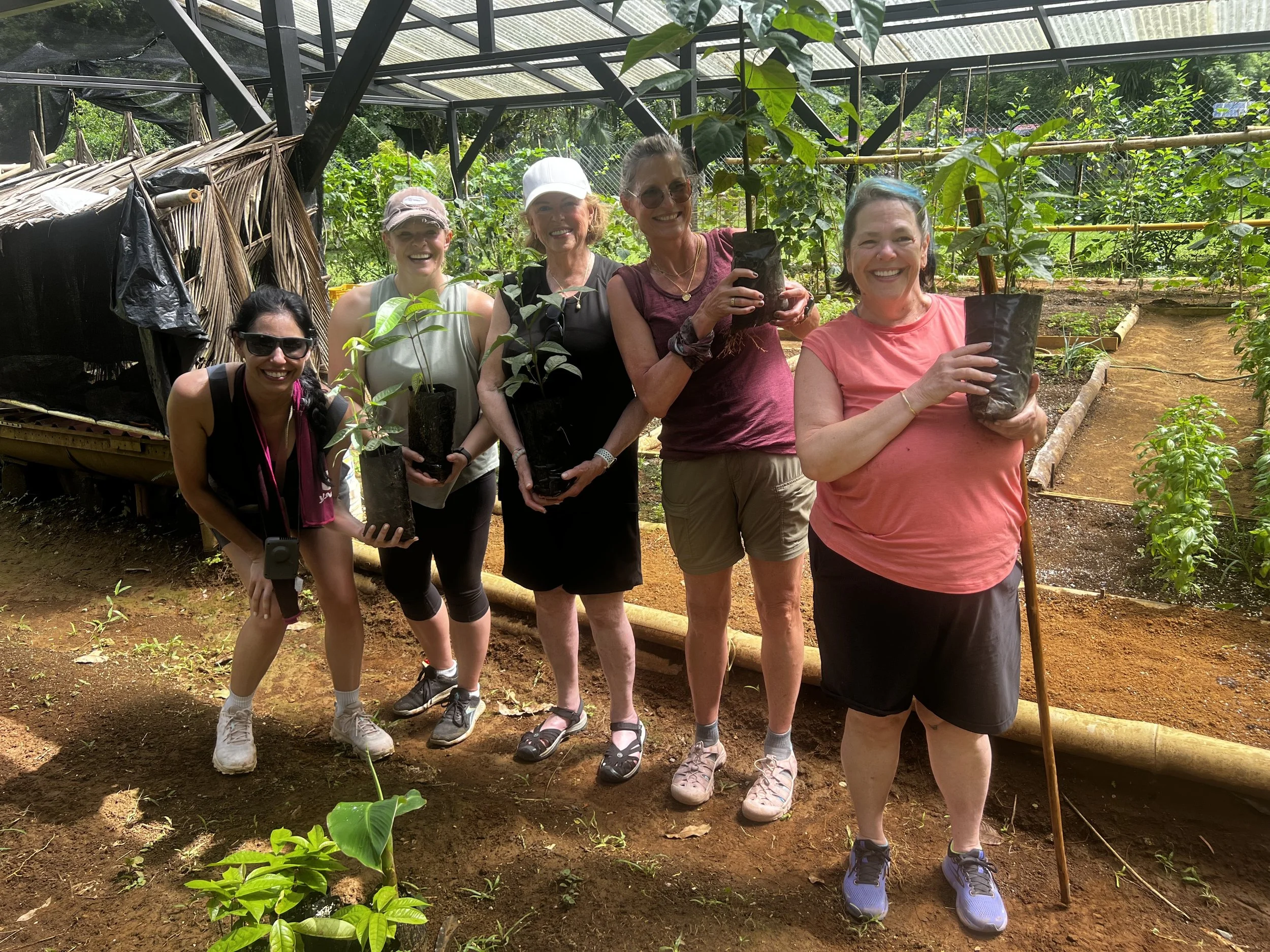 A group of six women smiling in a greenhouse, holding small potted trees, surrounded by lush green plants and garden structures.
