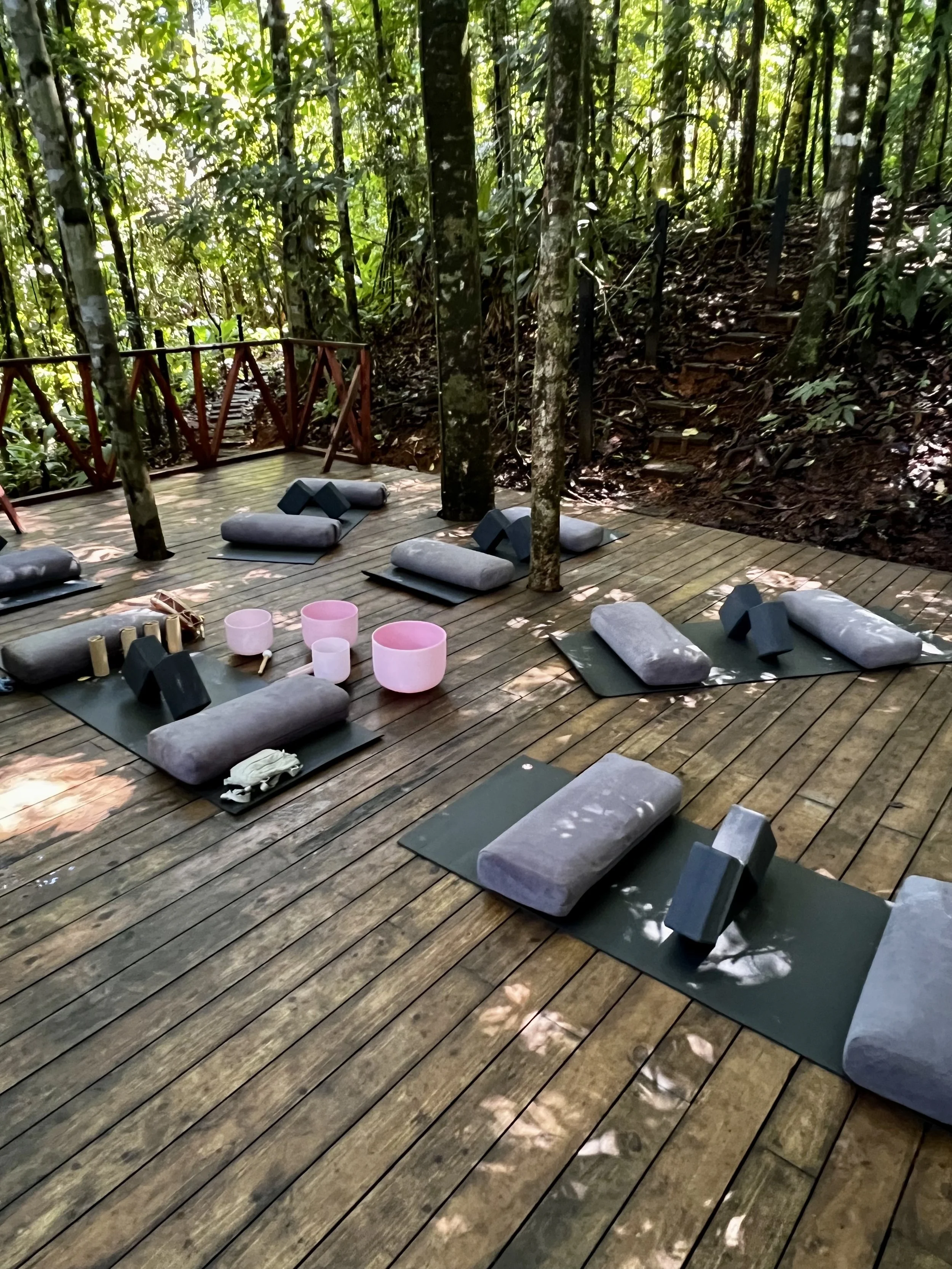 Outdoor sound bath and meditation setup with mats, cushions, and sound bowls on a wooden deck surrounded by trees in a rainforest.
