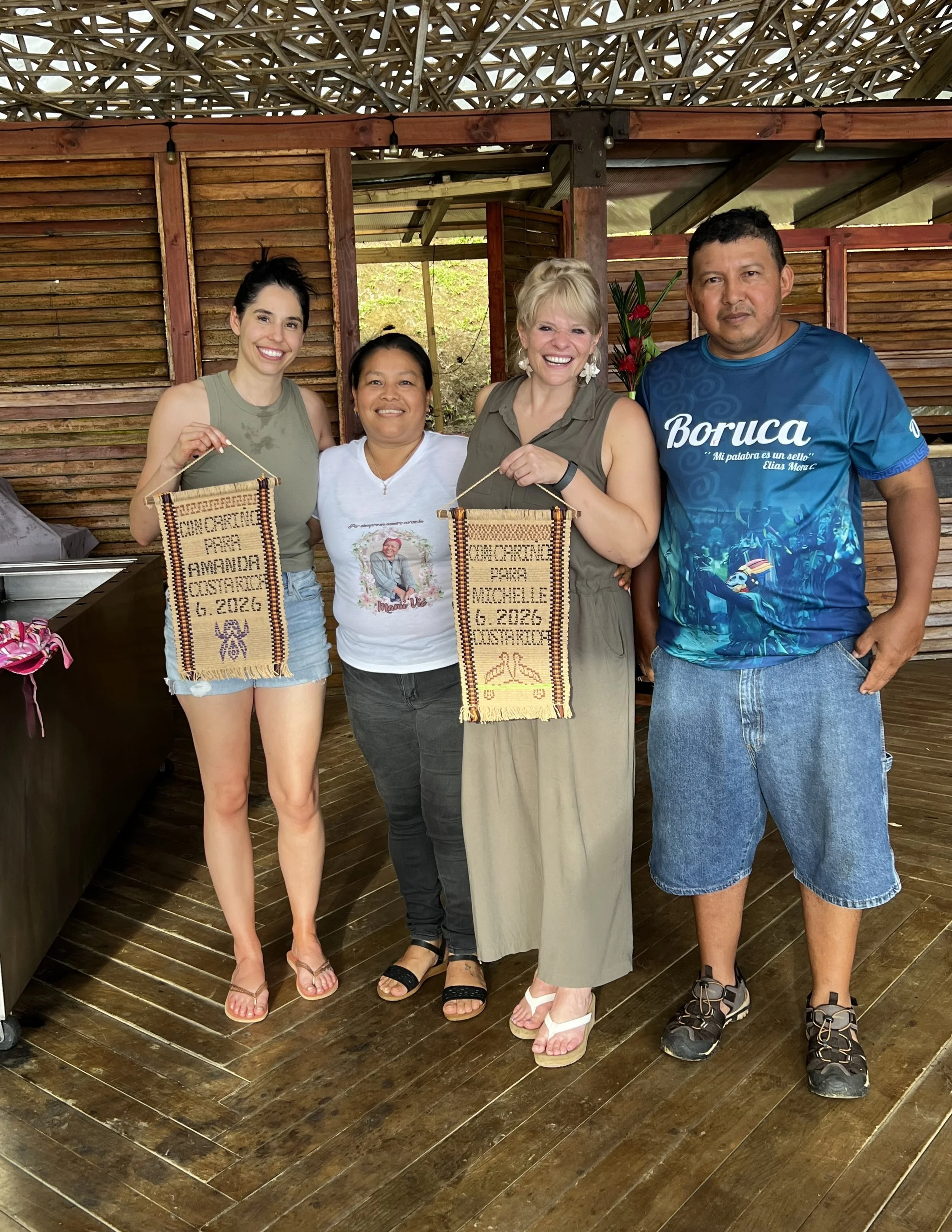 A group of four people standing together smiling and holding woven posters with text and symbols. The background is a wooden wall and the ceiling has a lattice design.