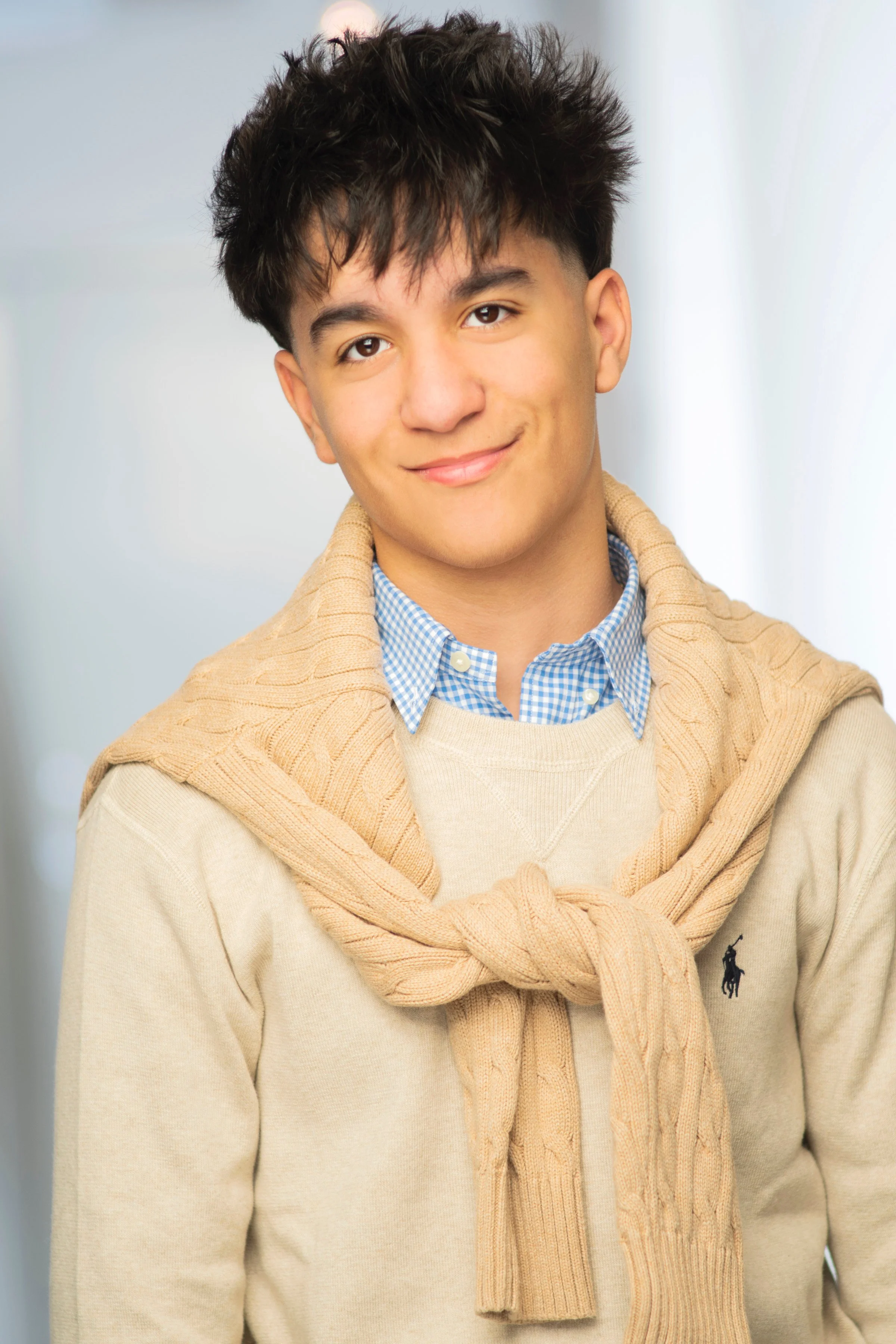 A young man with dark, messy hair smiling, wearing a beige sweater, a blue checkered shirt, and a knit sweater tied around his shoulders.