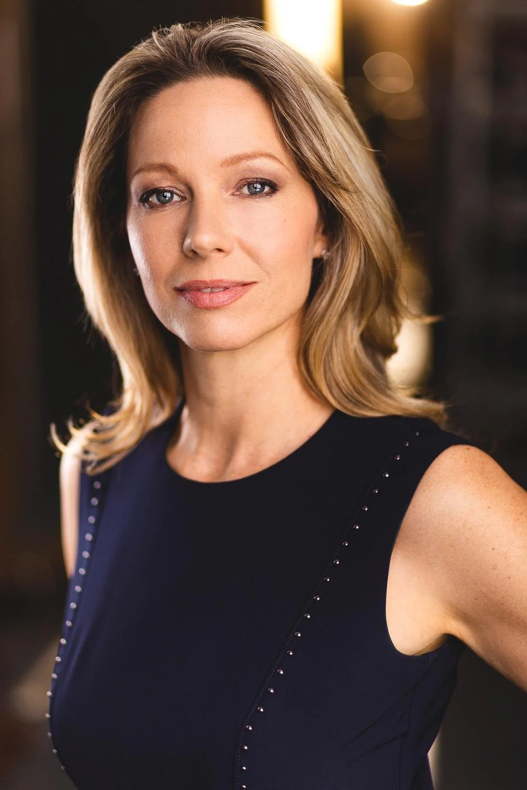 A woman with shoulder-length blonde hair and blue eyes, wearing a sleeveless black dress with small embellishments along the shoulder and side, standing indoors with warm lighting.