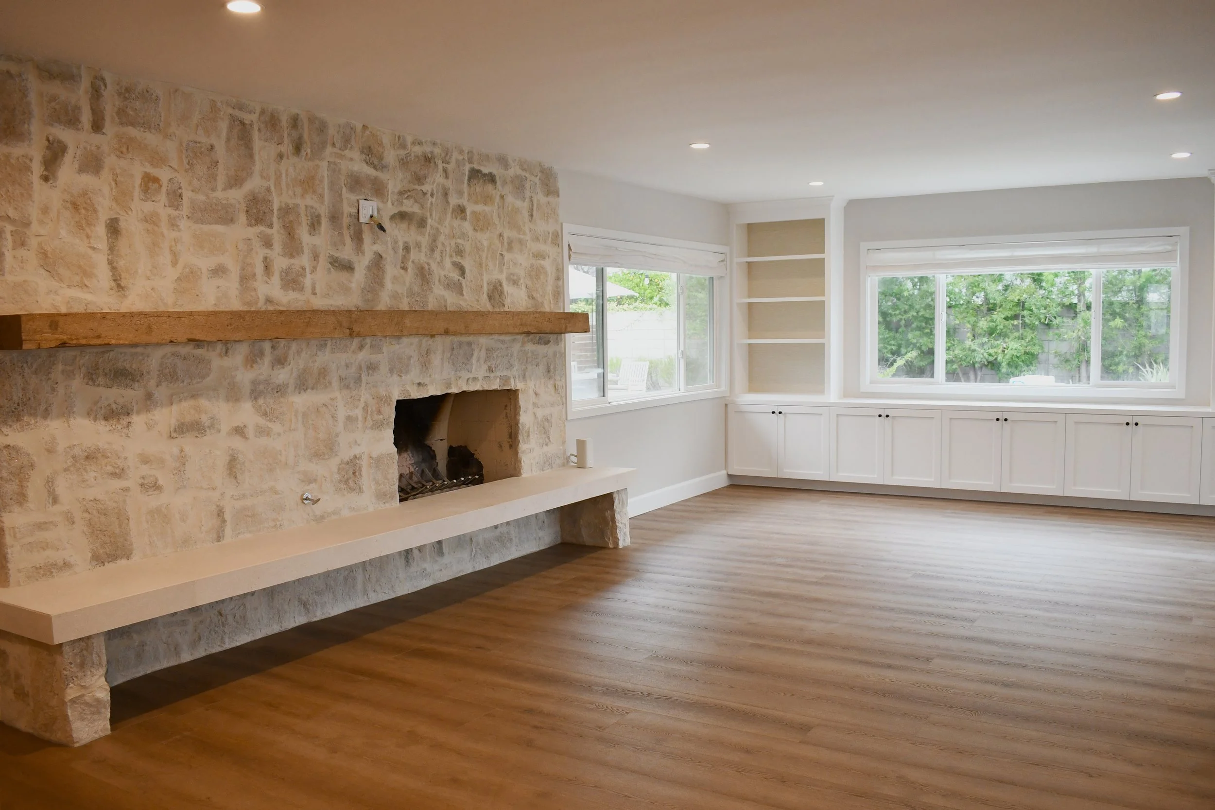 Empty living room with stone fireplace, wooden floor, large windows, and built-in shelves.