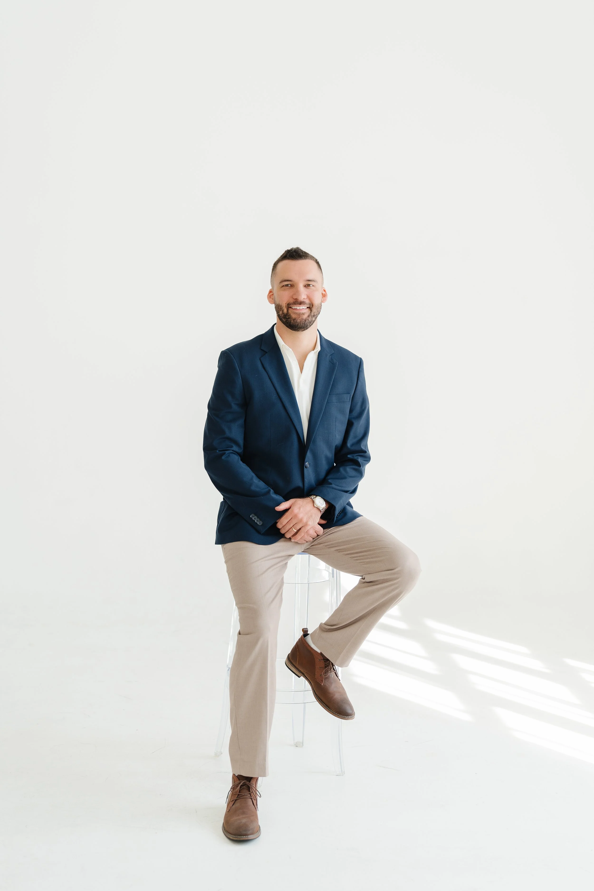 A smiling man with a beard is sitting on a clear chair against a plain white background. He's wearing a navy blazer, white dress shirt, beige pants, and brown boots, with one leg crossed over the other and hands resting on his knee.