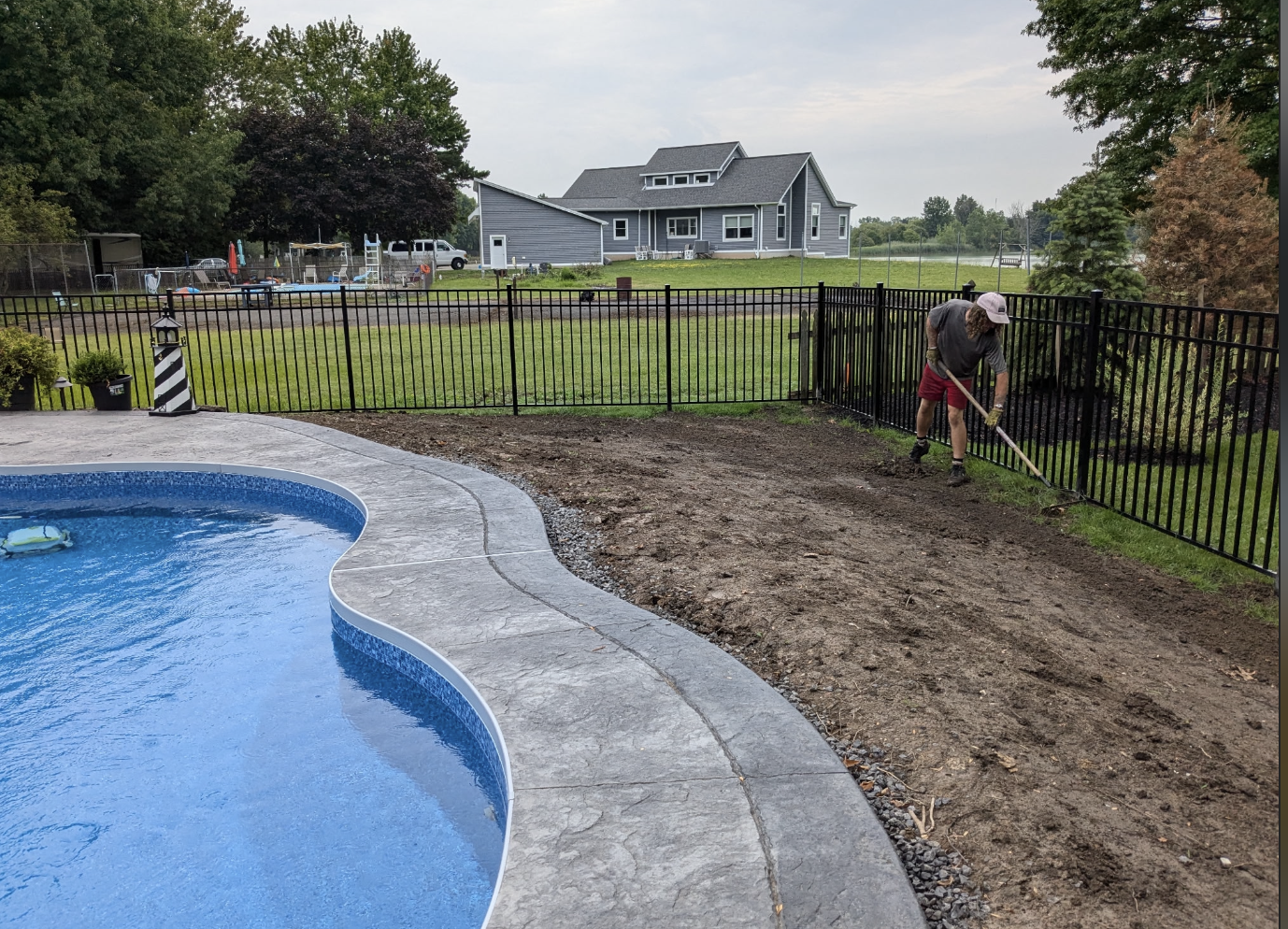 A person raking soil beside an outdoor swimming pool with a black metal fence surrounding the area. A residential house and garden are visible in the background.