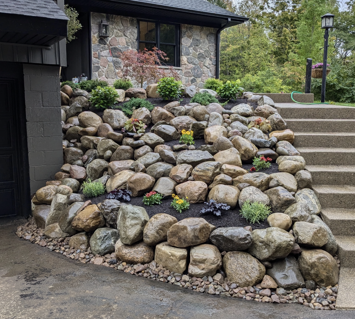 Stone landscaping with various plants and flowers on a sloped yard next to a house with a stone exterior and steps leading up to a lawn.