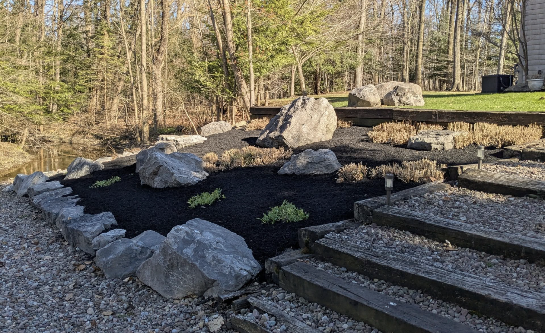 A landscaped backyard with large rocks, small plants, and a black mulch area, near the water and trees.