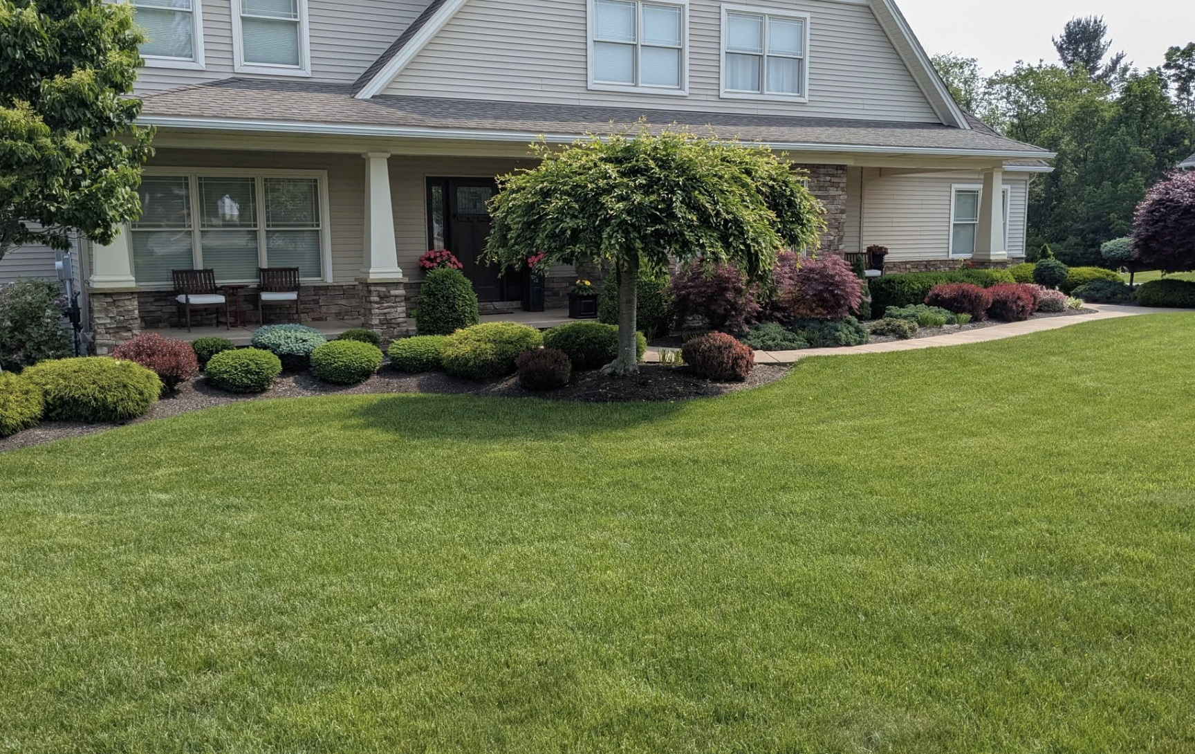Front yard of a house with neatly trimmed green lawn, various shrubs, small trees, and a porch with chairs and flowers.