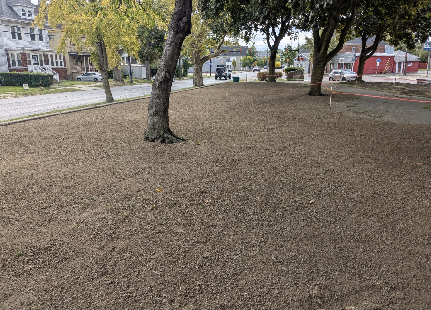 A park with freshly spread soil and several mature trees, with a street and residential houses in the background.
