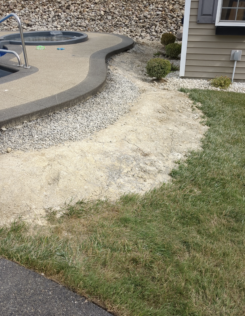 Outdoor pool area with a concrete walkway, gravel, grass lawn, and shrubbery near a house.