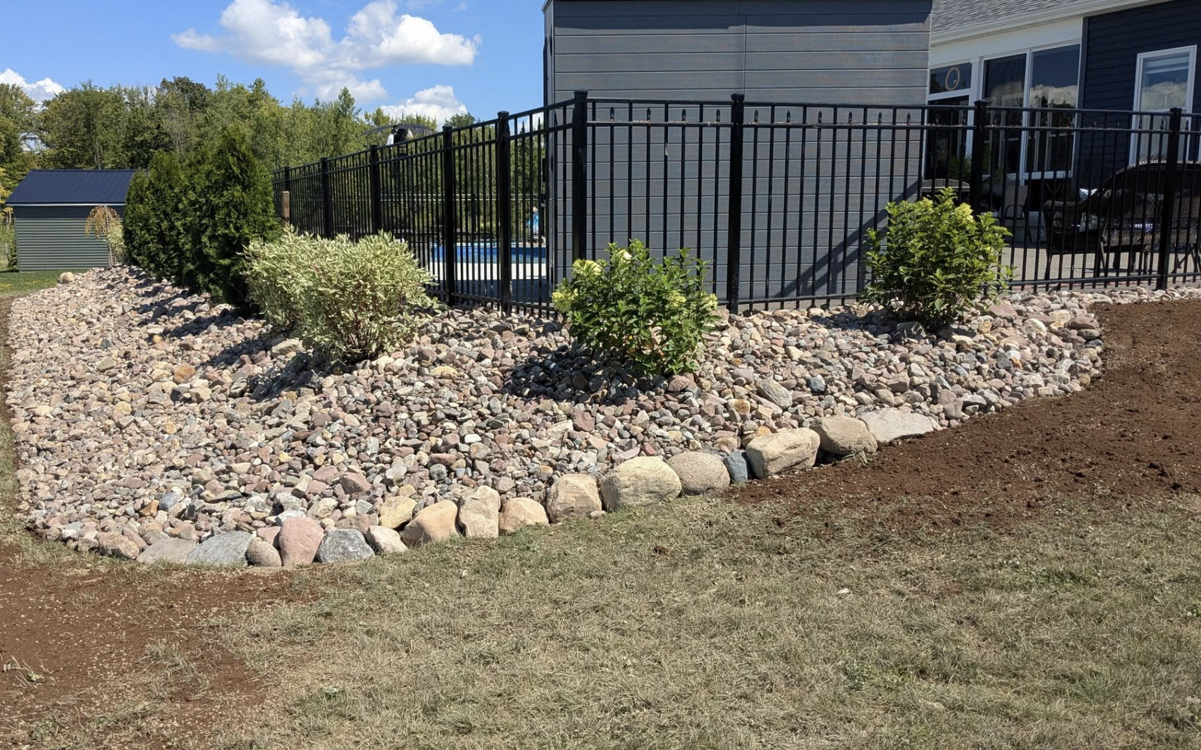 Landscape view of a backyard with a black metal fence, surrounding a blue swimming pool, and landscaped with rocks and bushes. There's a gray house and a small shed in the background, under a partly cloudy sky.