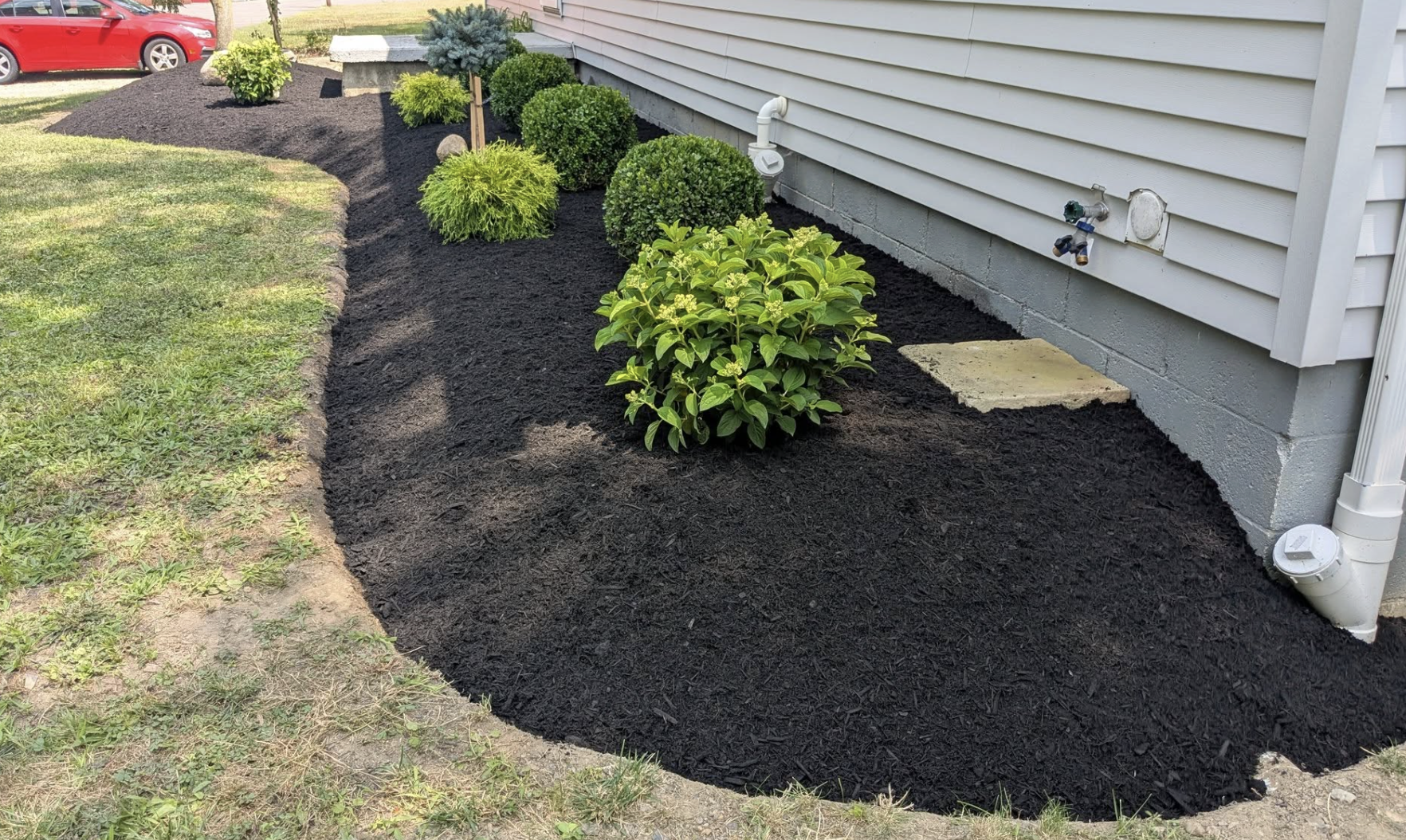 Freshly mulched garden bed with shrubs and bushes along the side of a house, with a red car parked nearby.