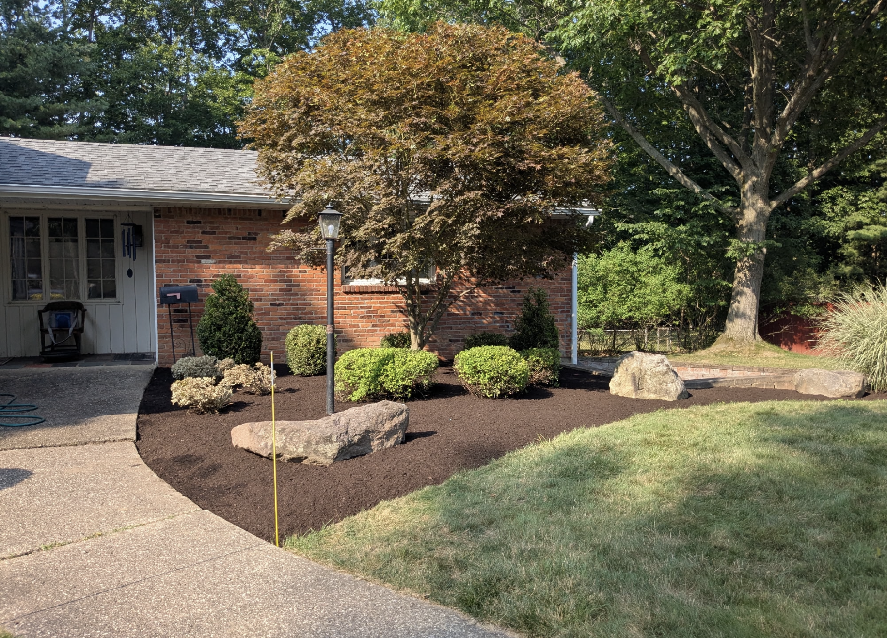 Residential front yard featuring a mulched garden bed with shrubs and trees, a brick house, a lamp post, large rocks, a concrete driveway, and a grassy lawn with trees in the background.