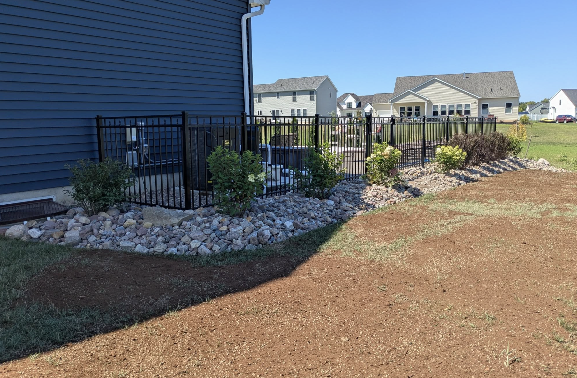 Backyard with a black metal fence, landscaped with rocks and plants, beside a house with blue siding, and neighboring houses visible in the background under a clear blue sky.