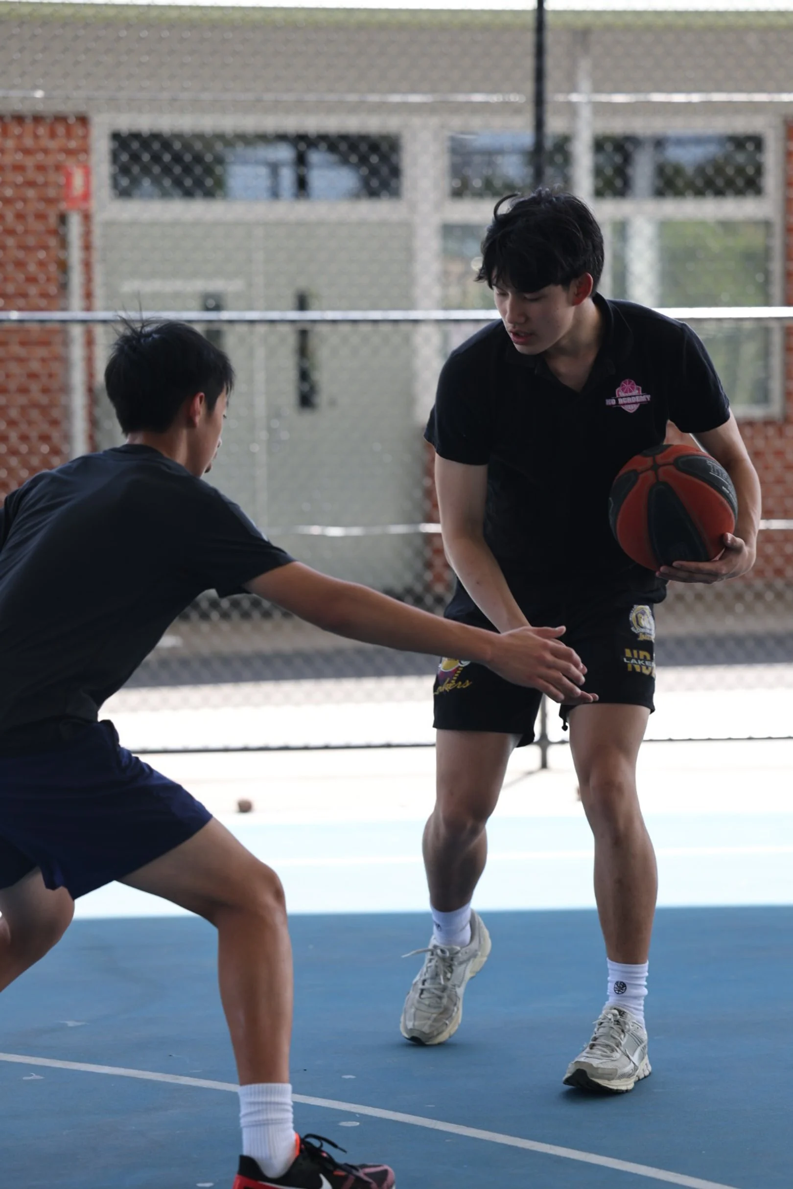 Two young boys playing basketball on an outdoor court near a chain-link fence, with one boy dribbling and the other defending.