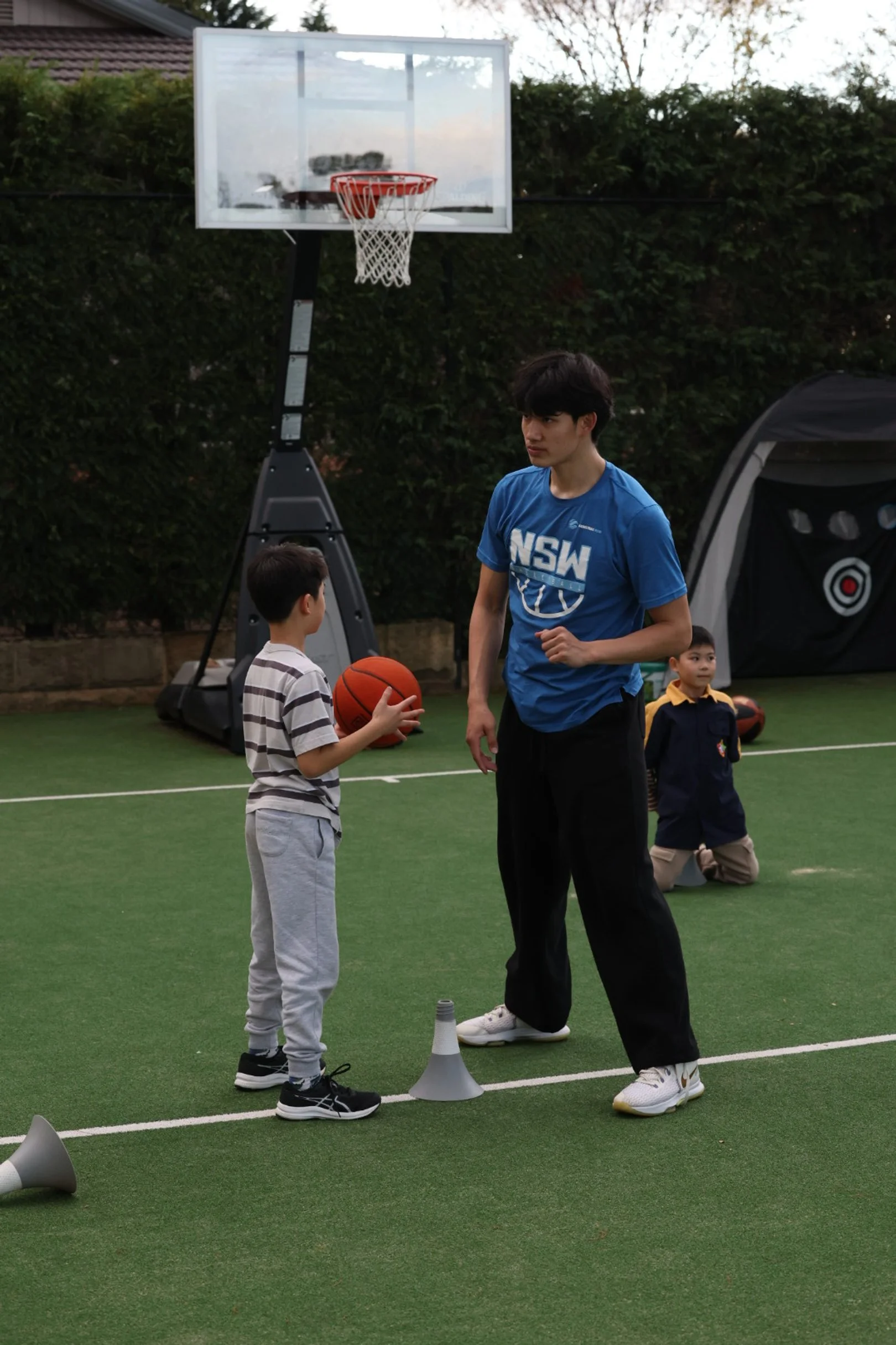 A young boy holding a basketball and talking to a coach on an outdoor basketball court, with children in the background, a basketball hoop, and green hedges.
