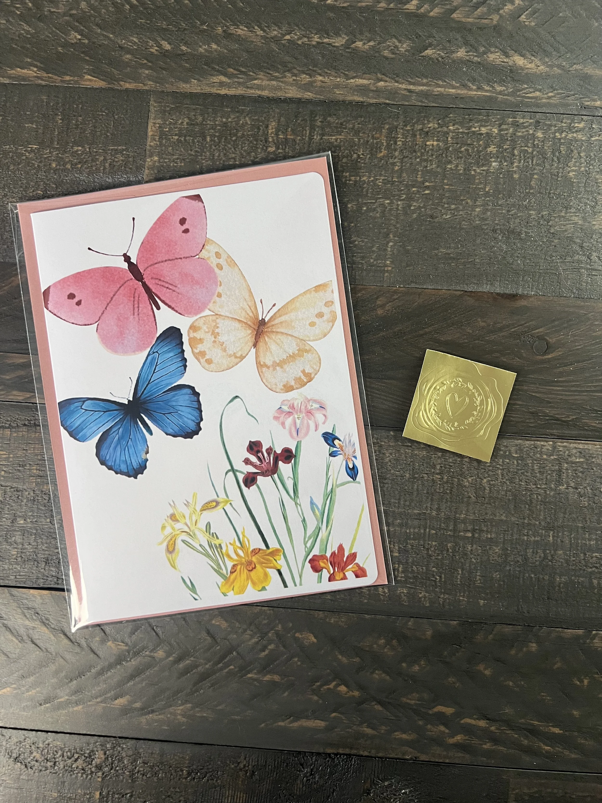 Packaged Mother’s Day card with butterfly and floral artwork, blush envelope, and gold wax seal sticker on wood surface