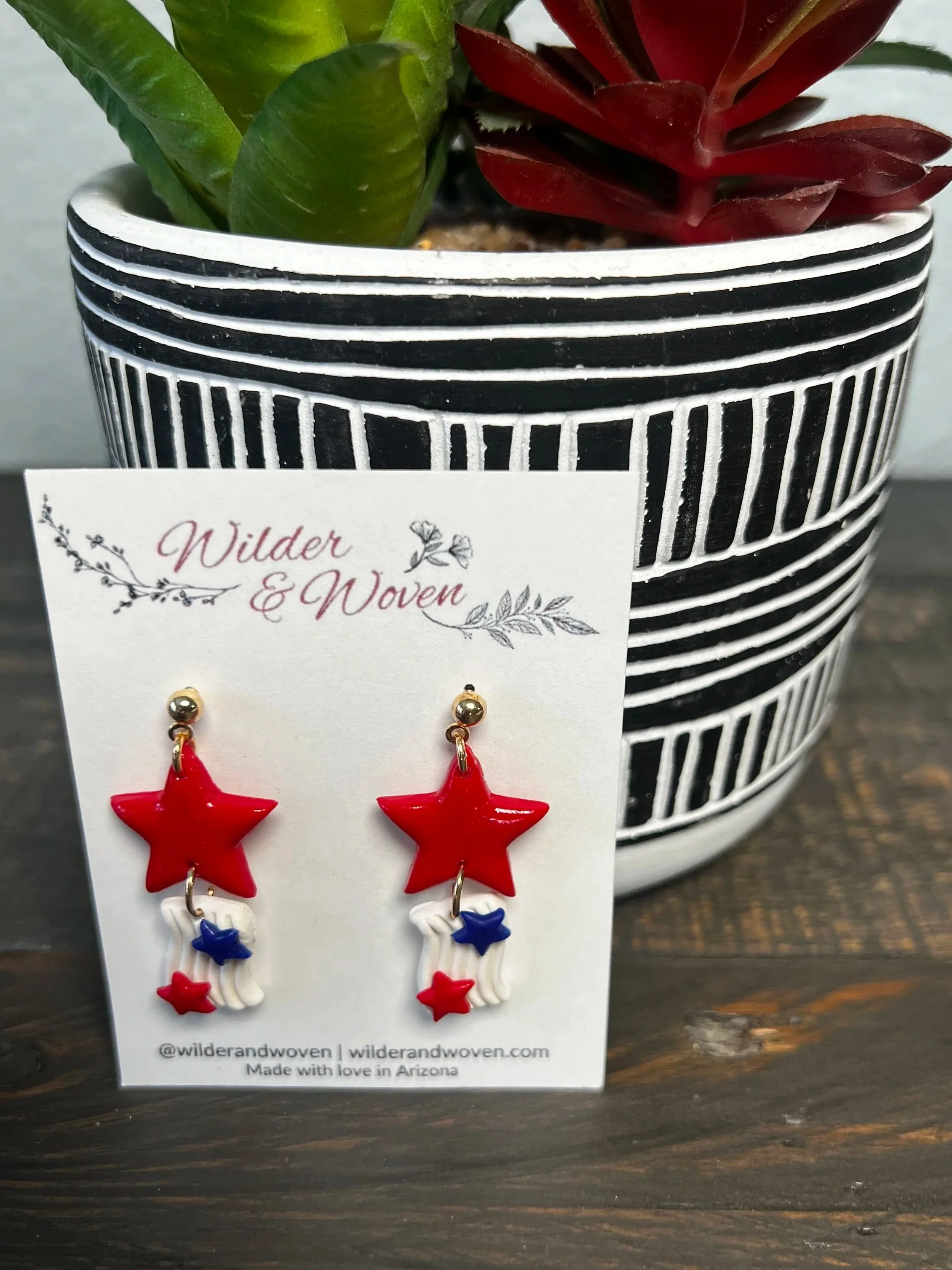Handmade patriotic polymer clay earrings featuring red stars with dangling red, white, and blue star accents suspended from gold stud posts on a Wilder & Woven display card.