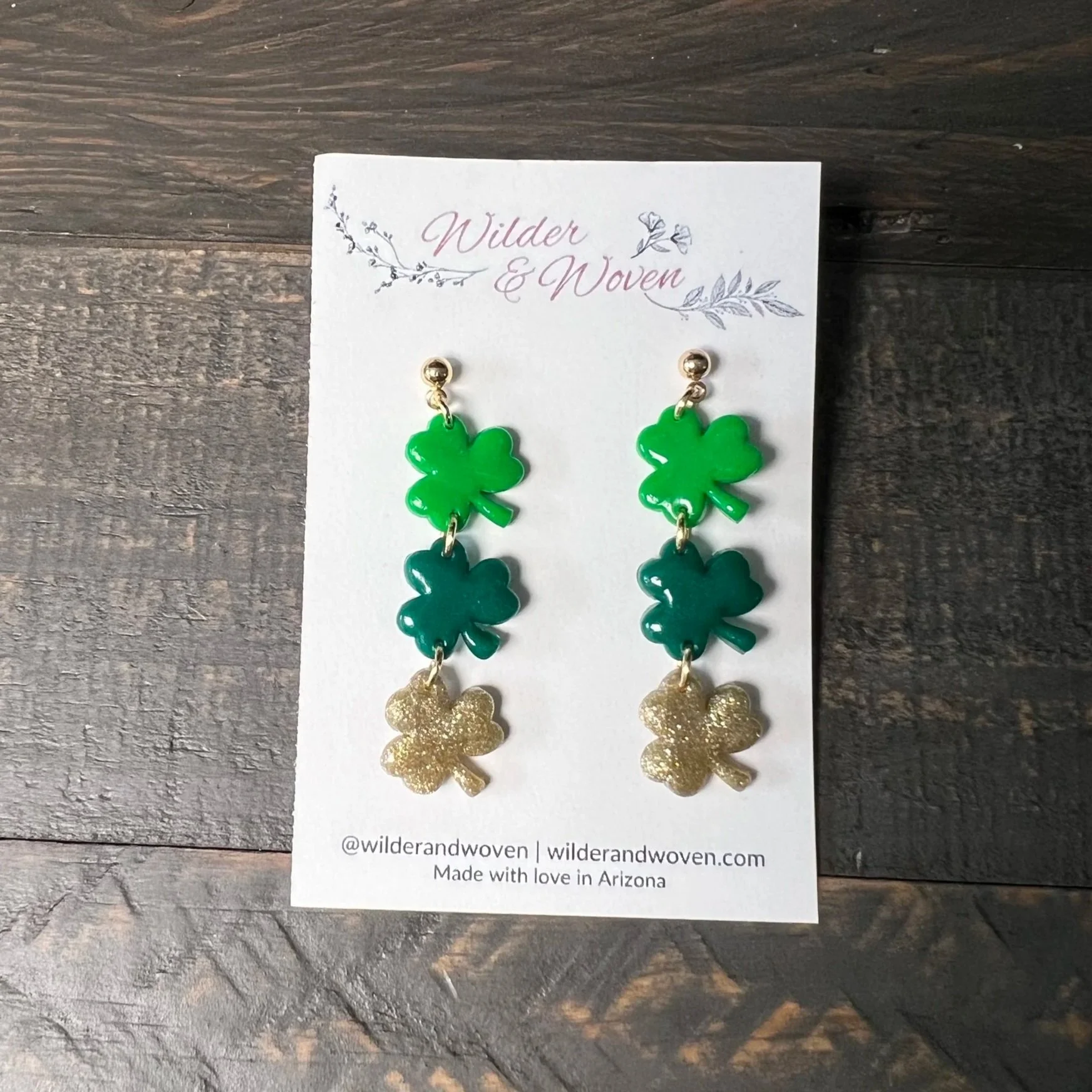 Handmade layered shamrock dangle earrings in green and gold polymer clay displayed on a Wilder & Woven earring card on a wooden table.