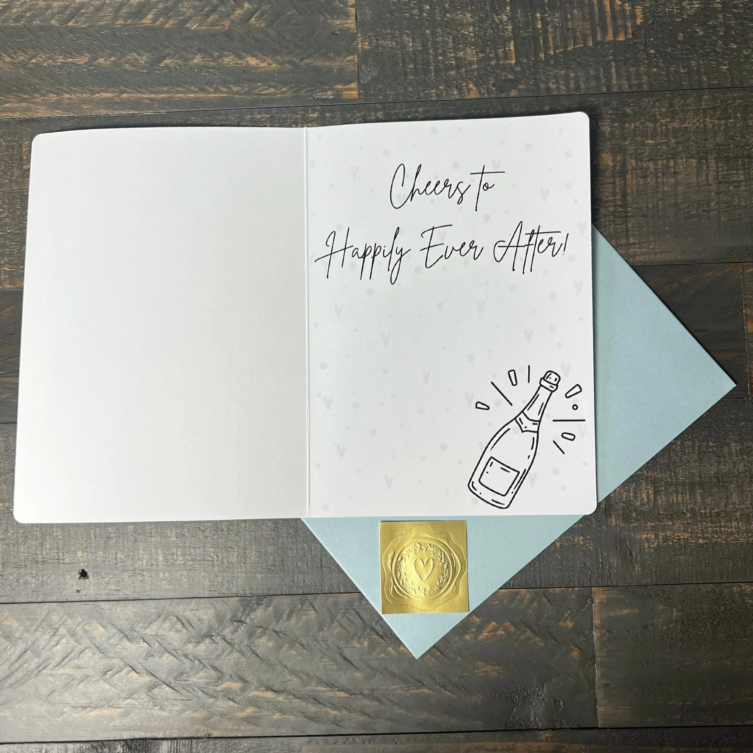 Open Cheers to Happily Ever After Wedding Card with coordinating soft blue envelope and gold wax seal style sticker.