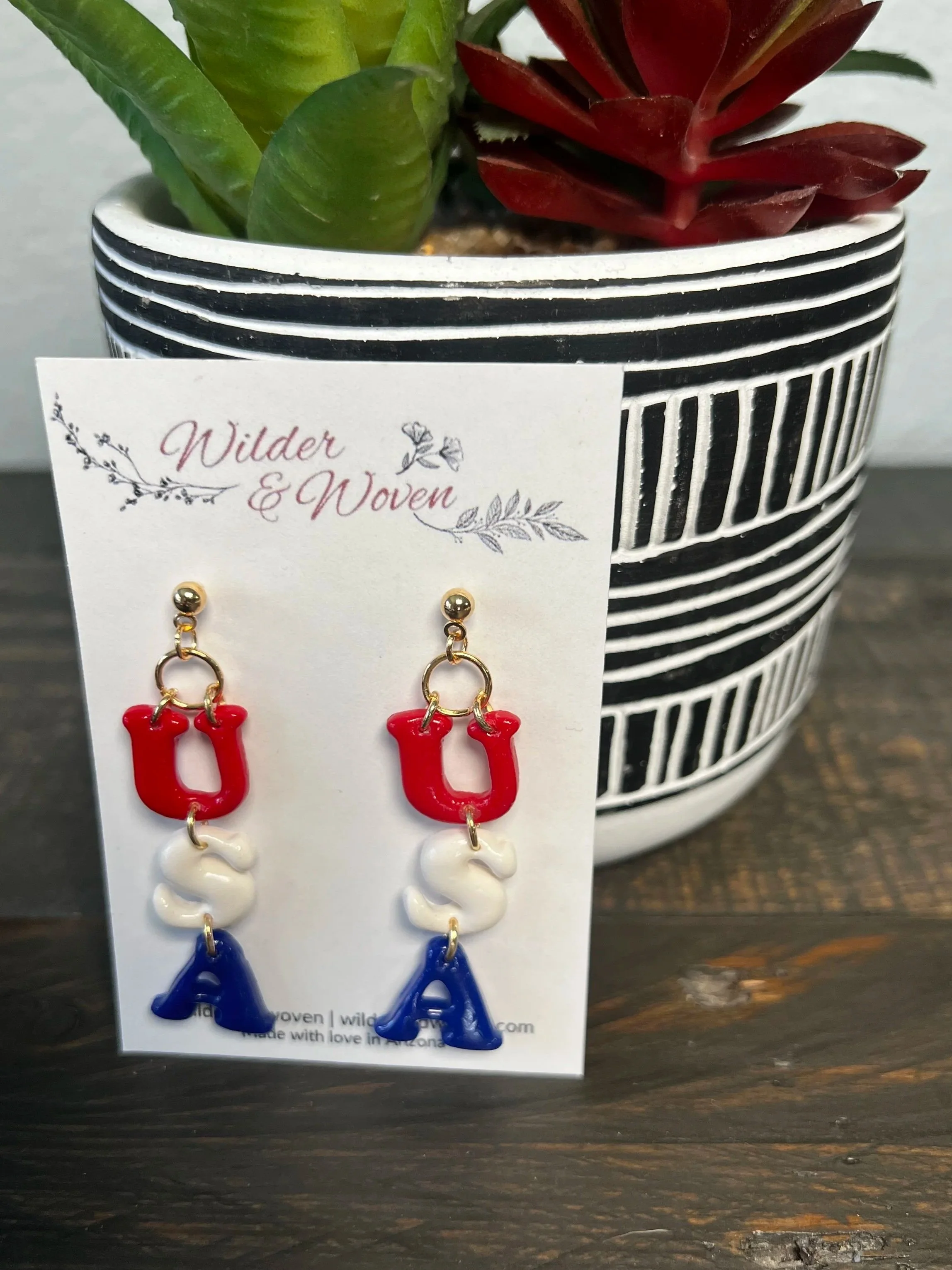 Handmade polymer clay USA letter earrings featuring red U, white S, and blue A charms hanging from gold stud posts on a Wilder & Woven display card.