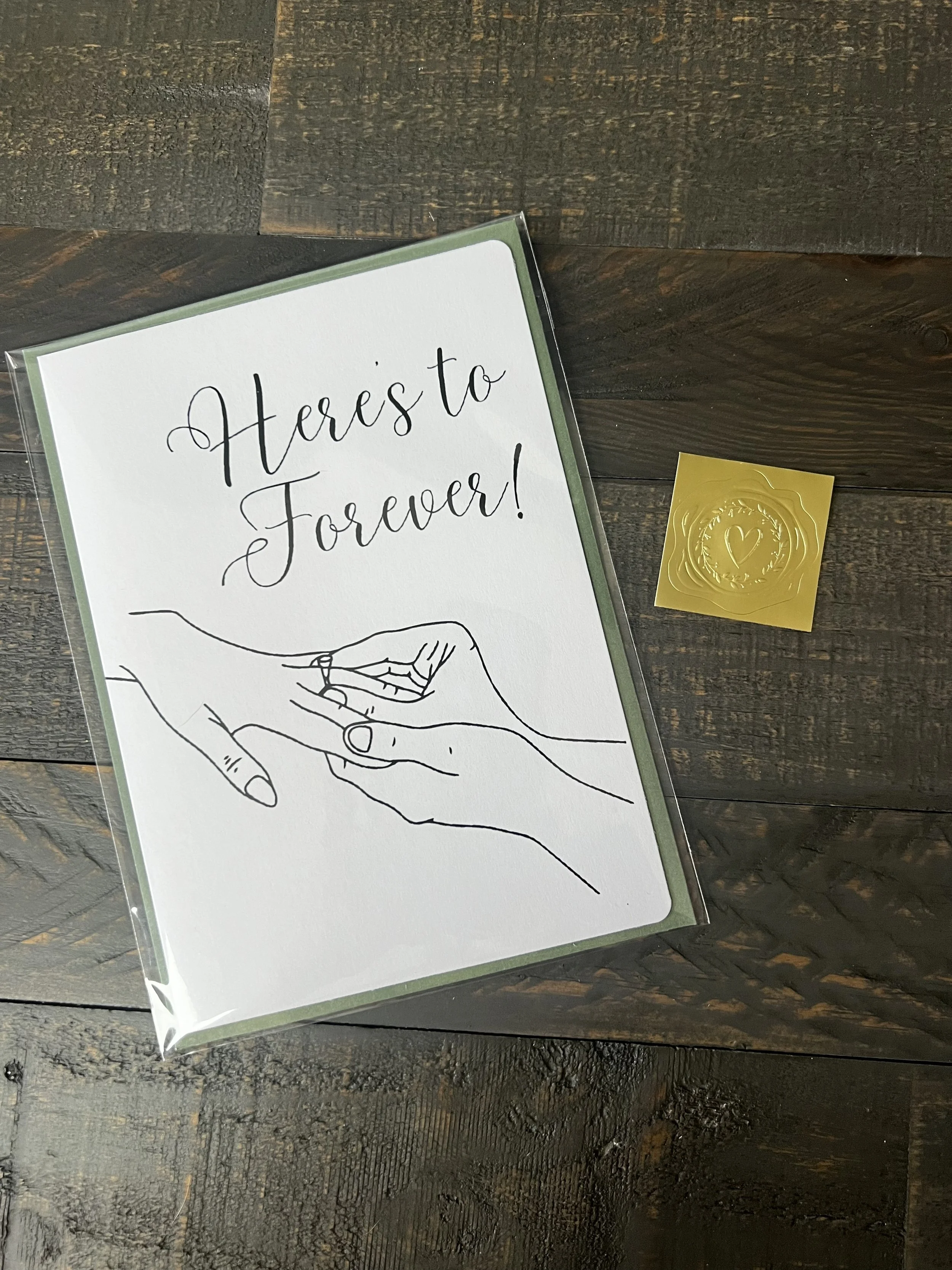 Styled flat lay of Here's to Forever engagement card with coordinating sage green envelope and gold wax seal style sticker.