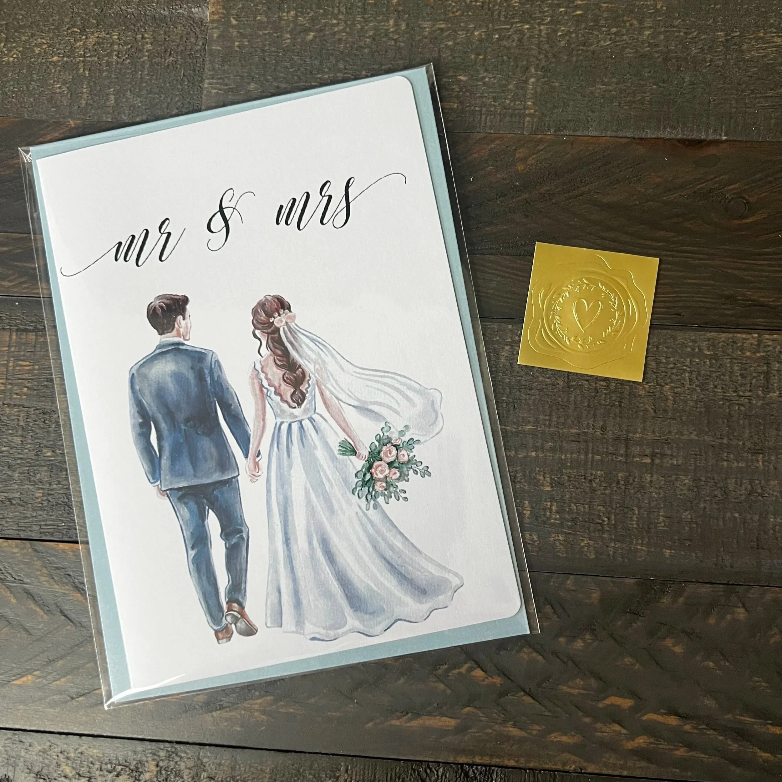 Styled flat lay of Mr. & Mrs. wedding greeting card featuring a brunette bride with coordinating soft blue envelope and gold wax seal style sticker.