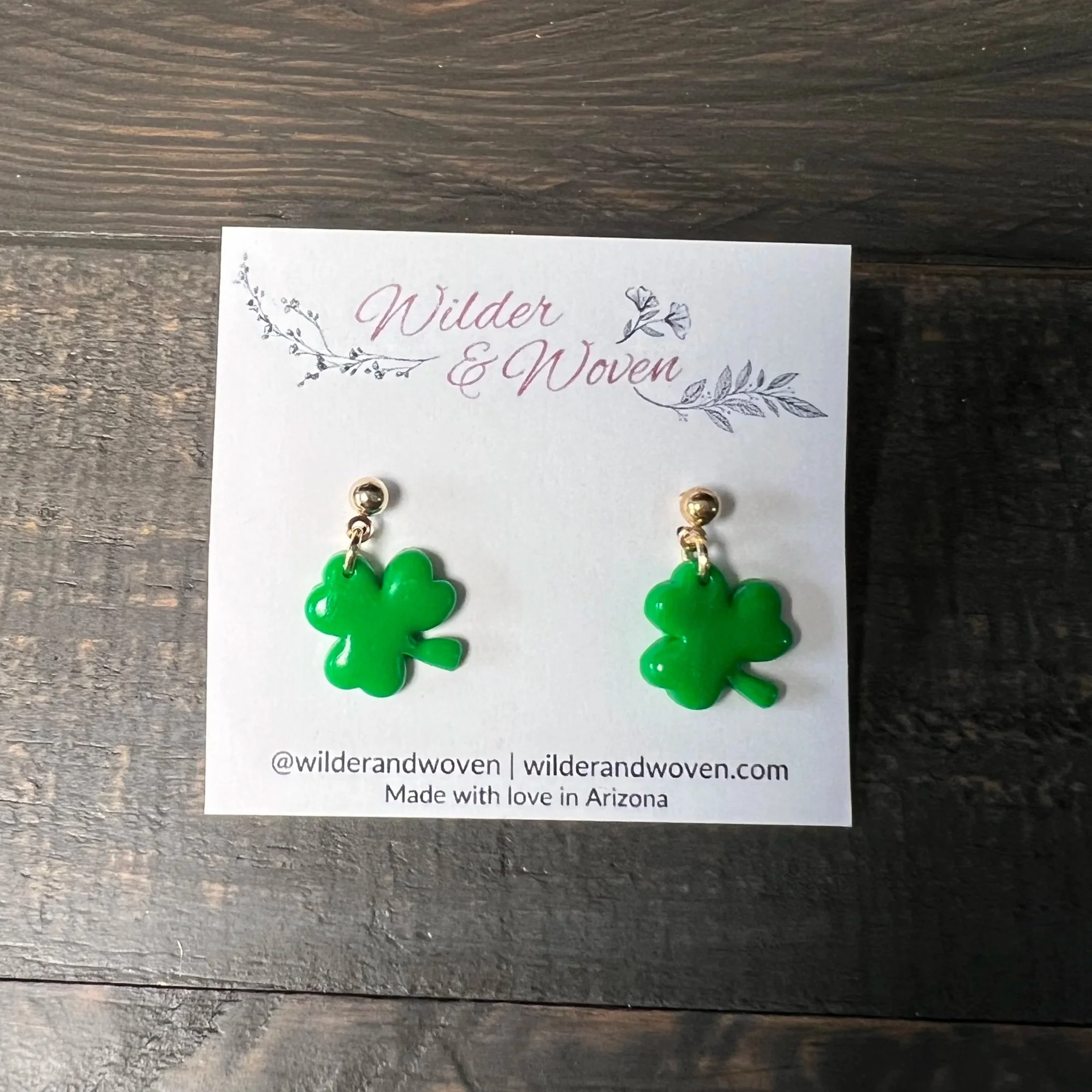 Handmade emerald green shamrock stud earrings displayed on a Wilder & Woven jewelry card on a wooden table.