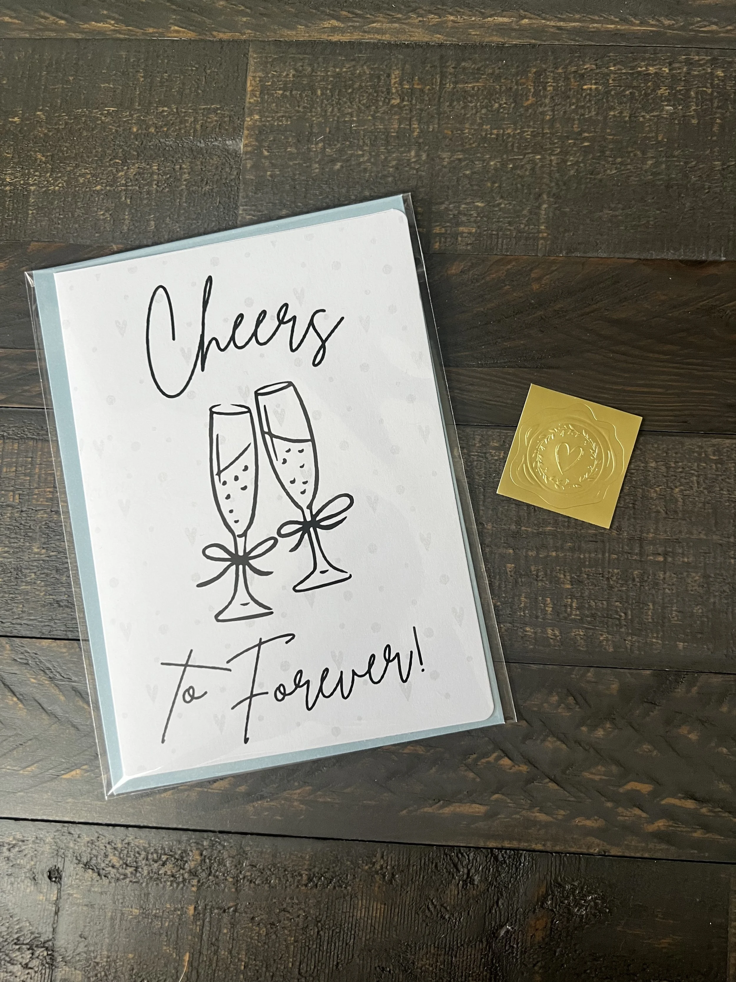 Styled flat lay of Cheers to Forever engagement card with coordinating soft blue envelope and gold wax seal style sticker.