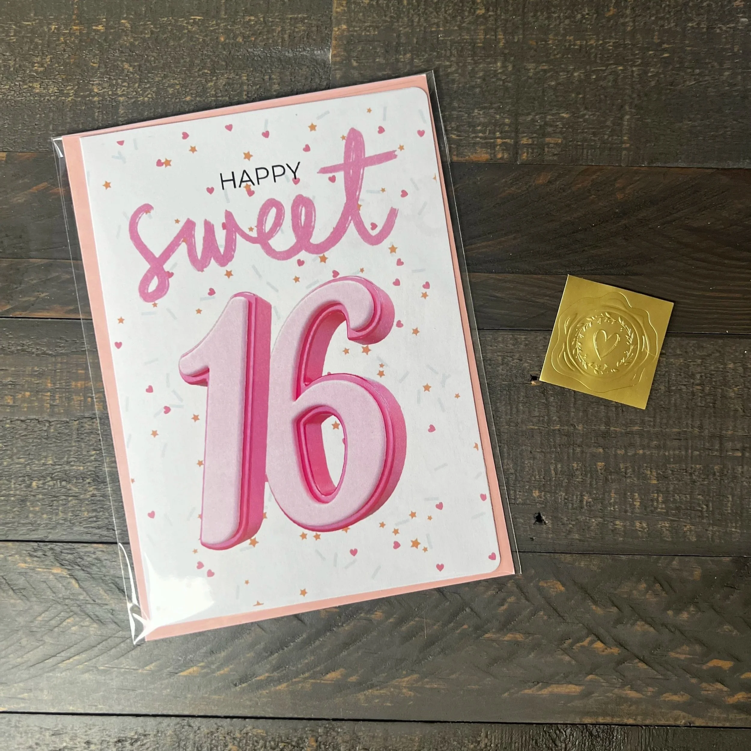 Styled flat lay of Sweet 16 greeting card with coordinating pink envelope and gold wax seal style sticker.