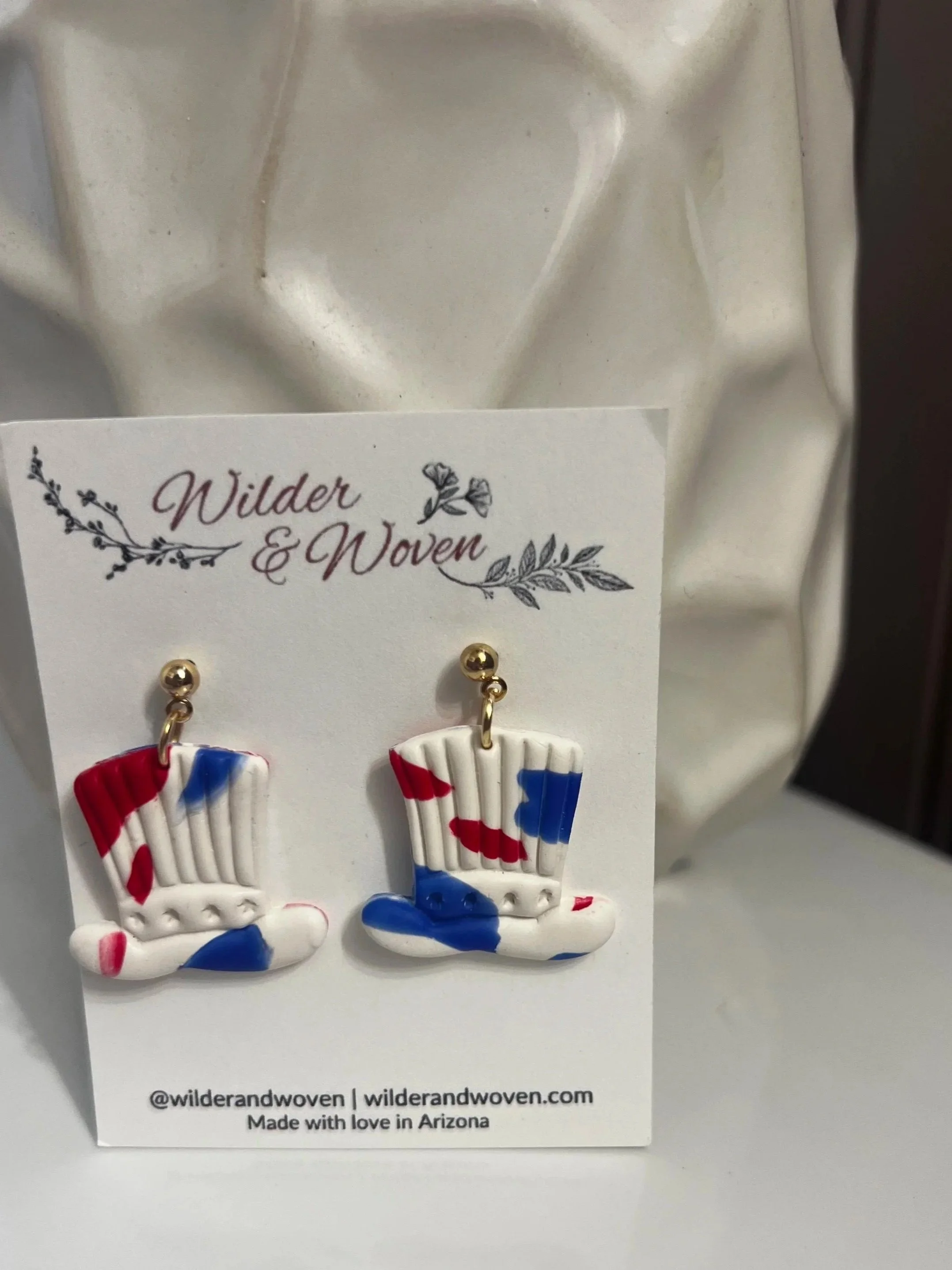 Handmade red, white, and blue polymer clay top hat earrings inspired by Uncle Sam, hanging from gold stud posts on a Wilder & Woven display card.
