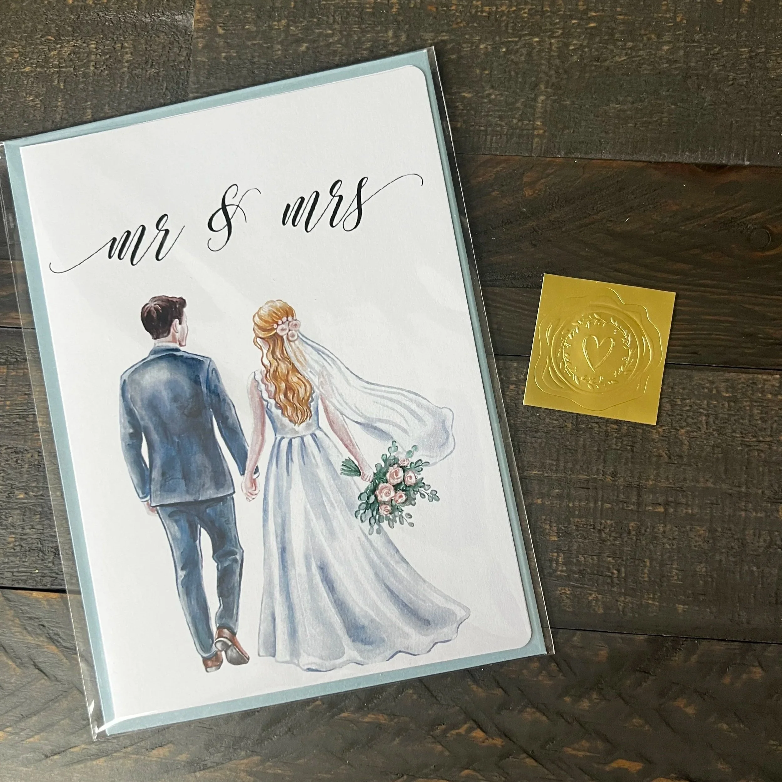 Styled flat lay of Mr. & Mrs. wedding greeting card featuring a blonde bride with coordinating soft blue envelope and gold wax seal style sticker.