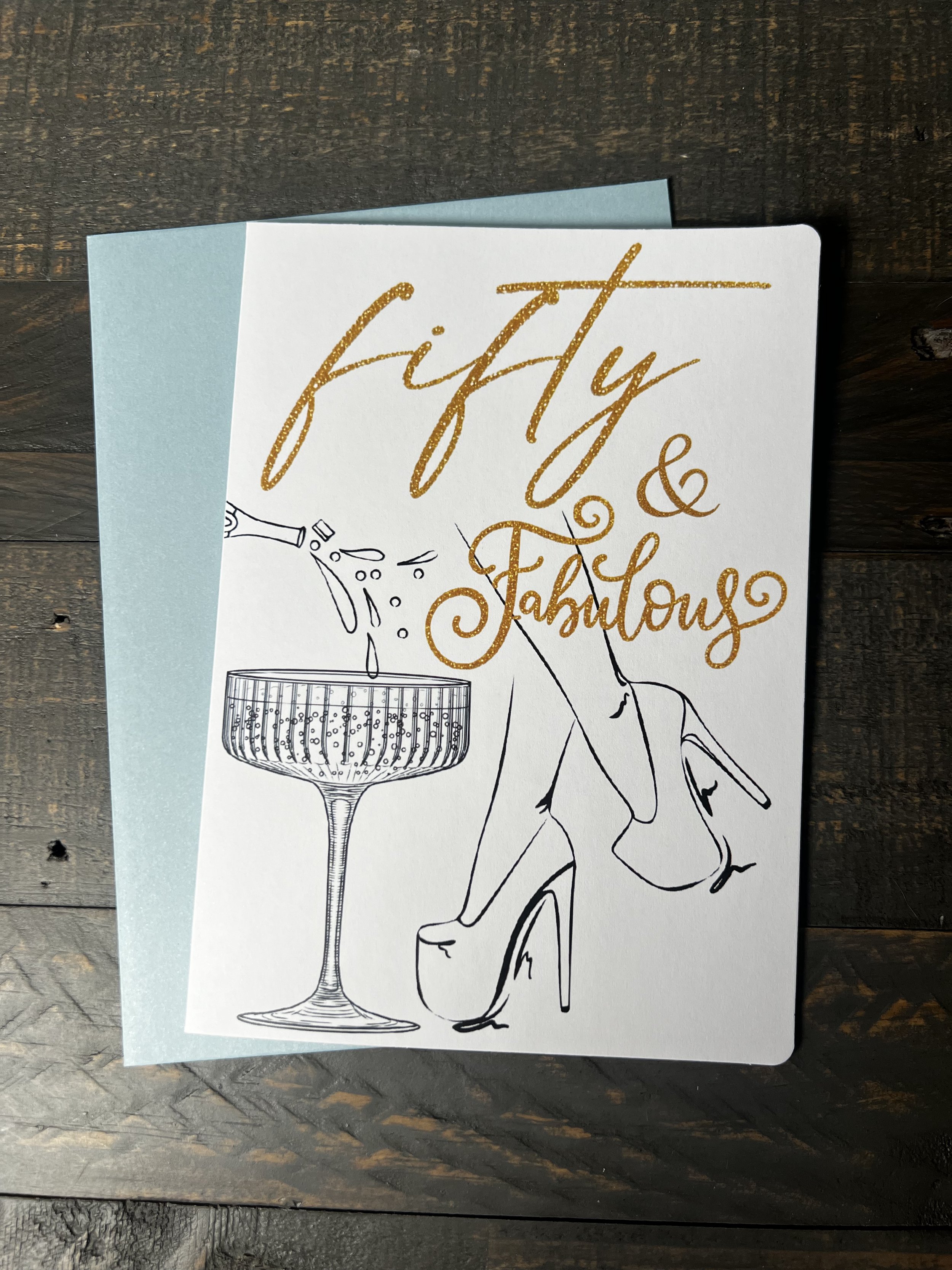 Fifty & Fabulous birthday greeting card featuring a champagne glass, elegant heels illustration, and coordinating soft blue envelope.