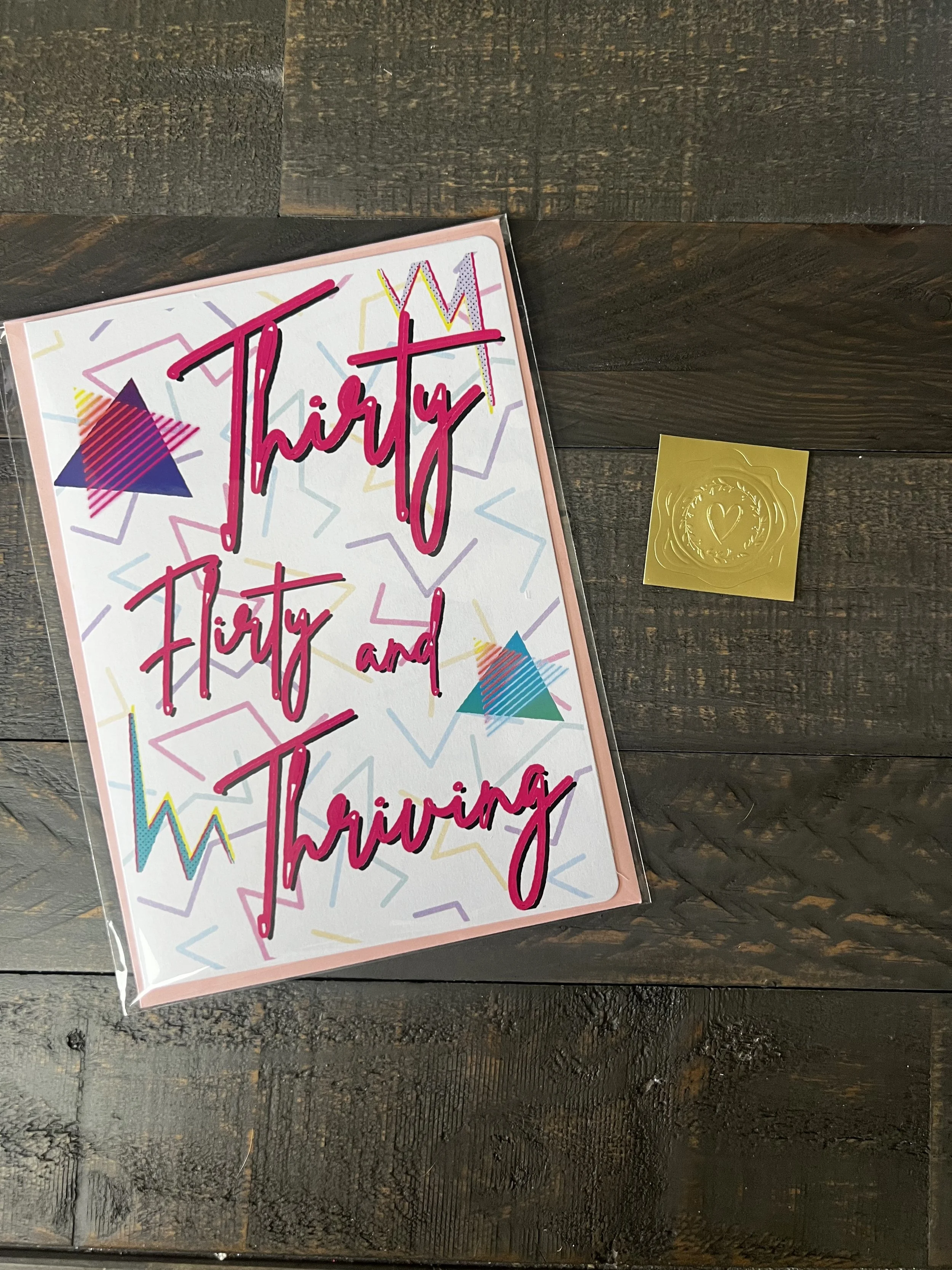Styled flat lay of Thirty, Flirty & Thriving greeting card with coordinating pink envelope and gold wax seal style sticker.