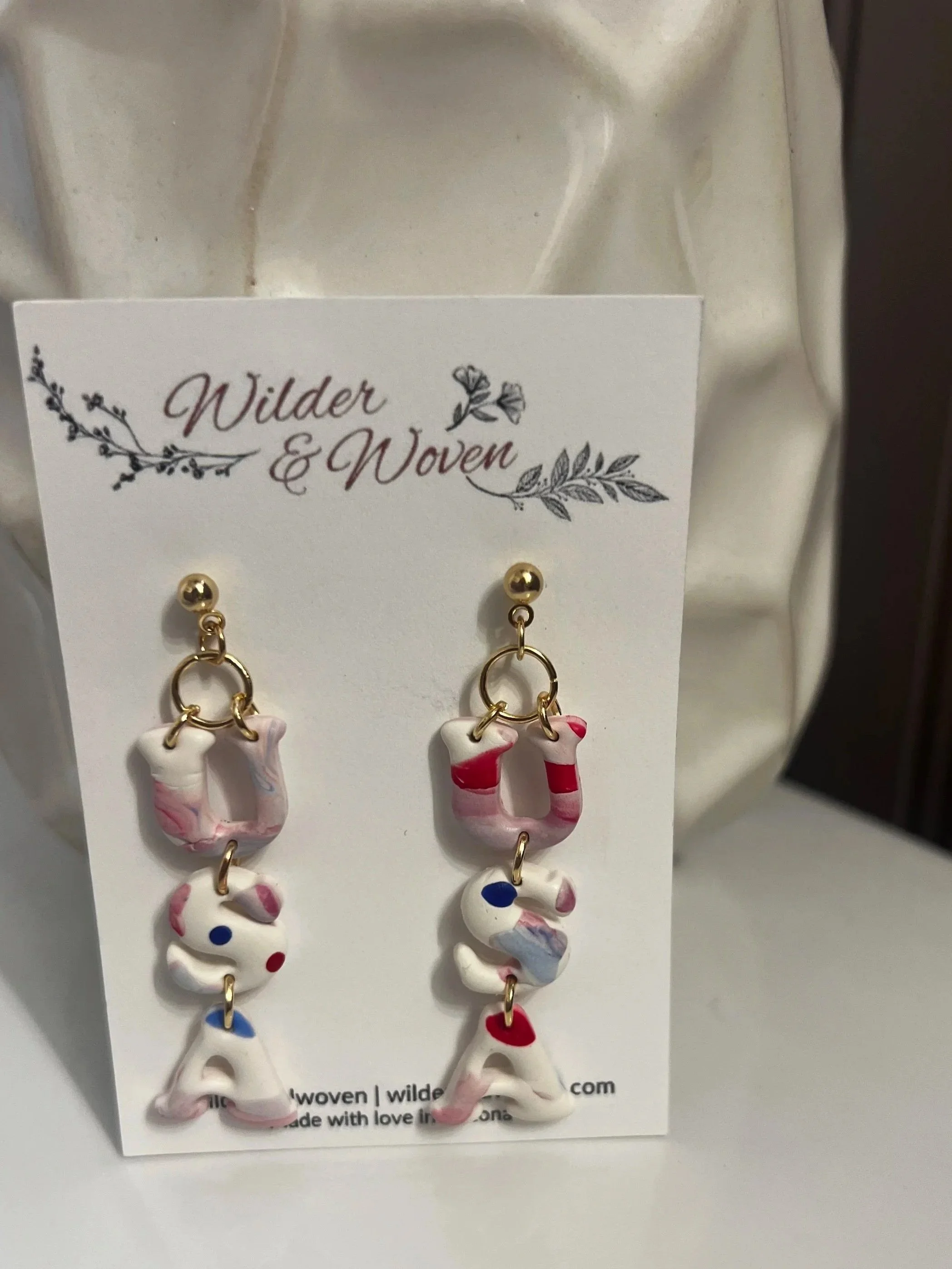 Handmade polymer clay USA letter earrings featuring blush pink marbling and patriotic red, white, and blue accents hanging from gold stud posts on a Wilder & Woven display card.