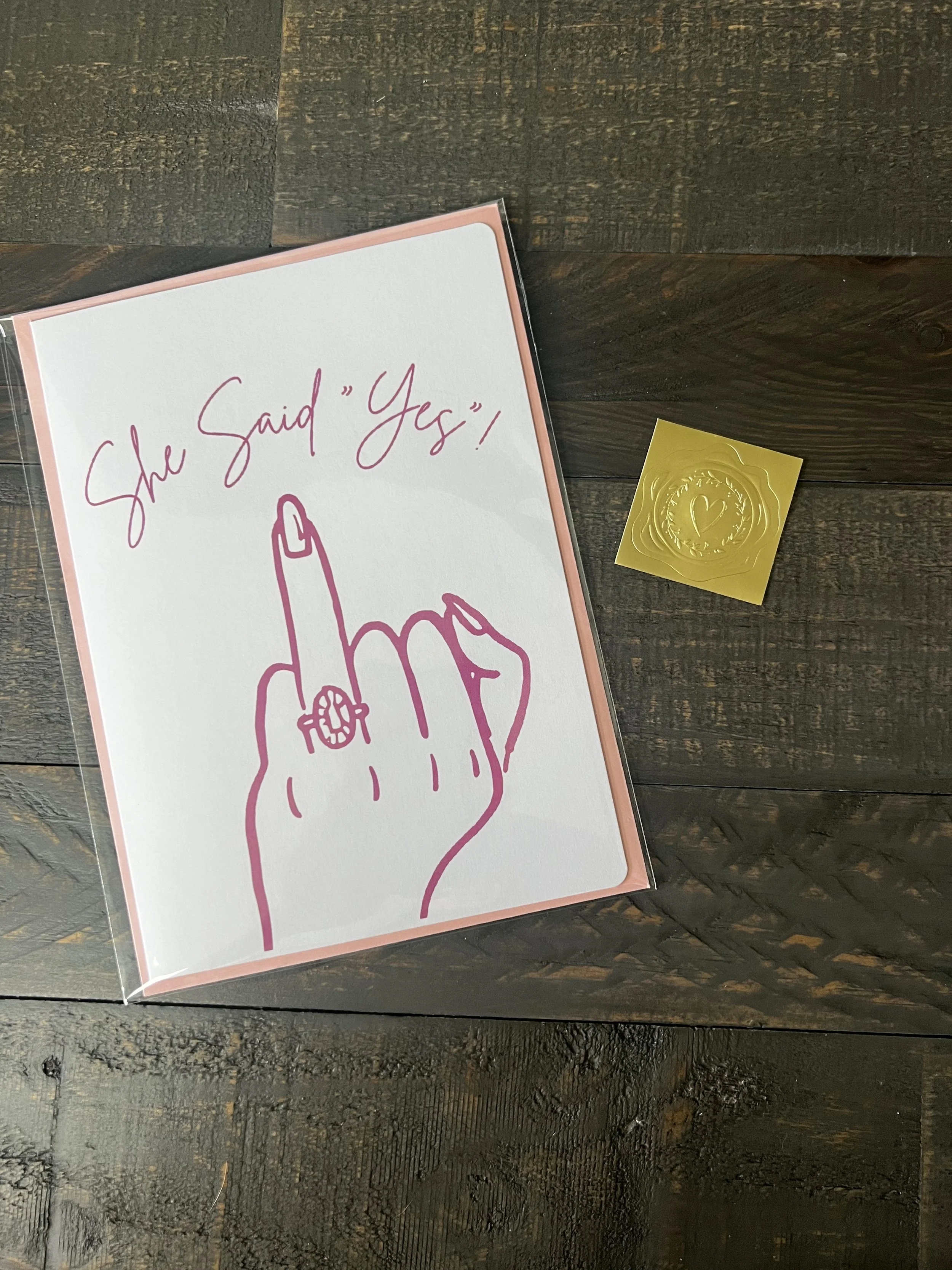 Styled flat lay of She Said Yes! engagement card with coordinating blush pink envelope and gold wax seal style sticker.