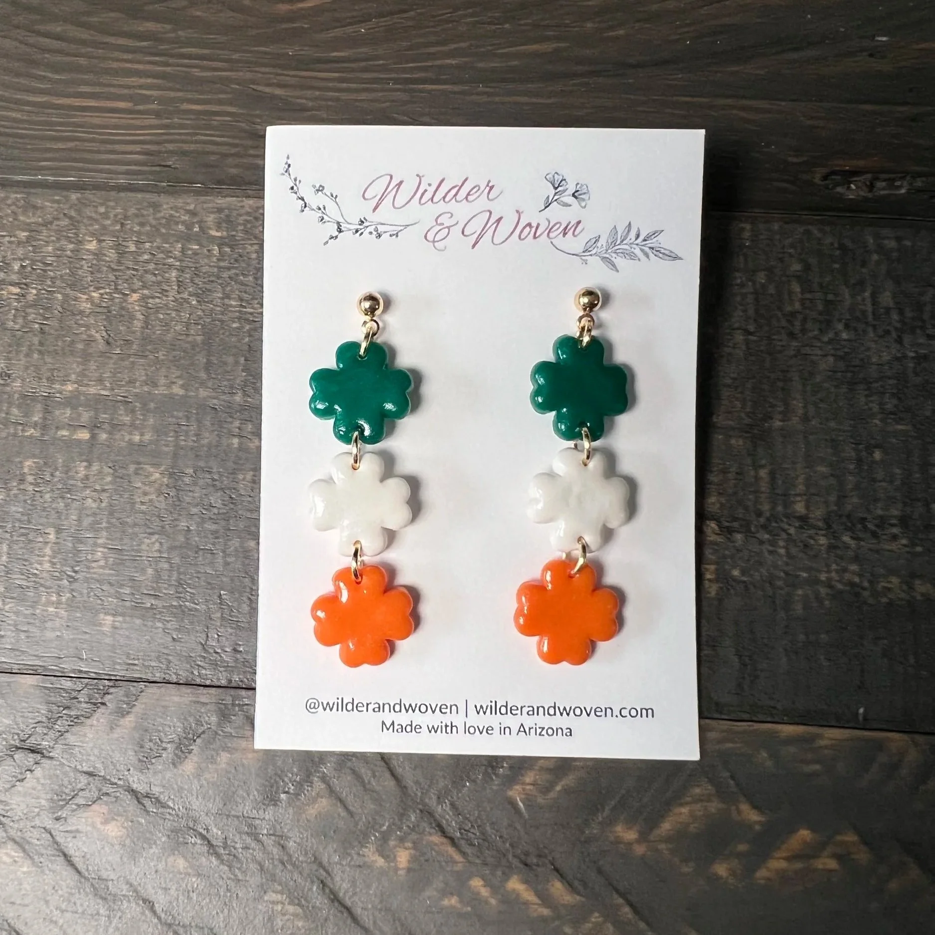 Handmade green, white, and orange shamrock dangle earrings displayed on a Wilder & Woven earring card on a wooden table.