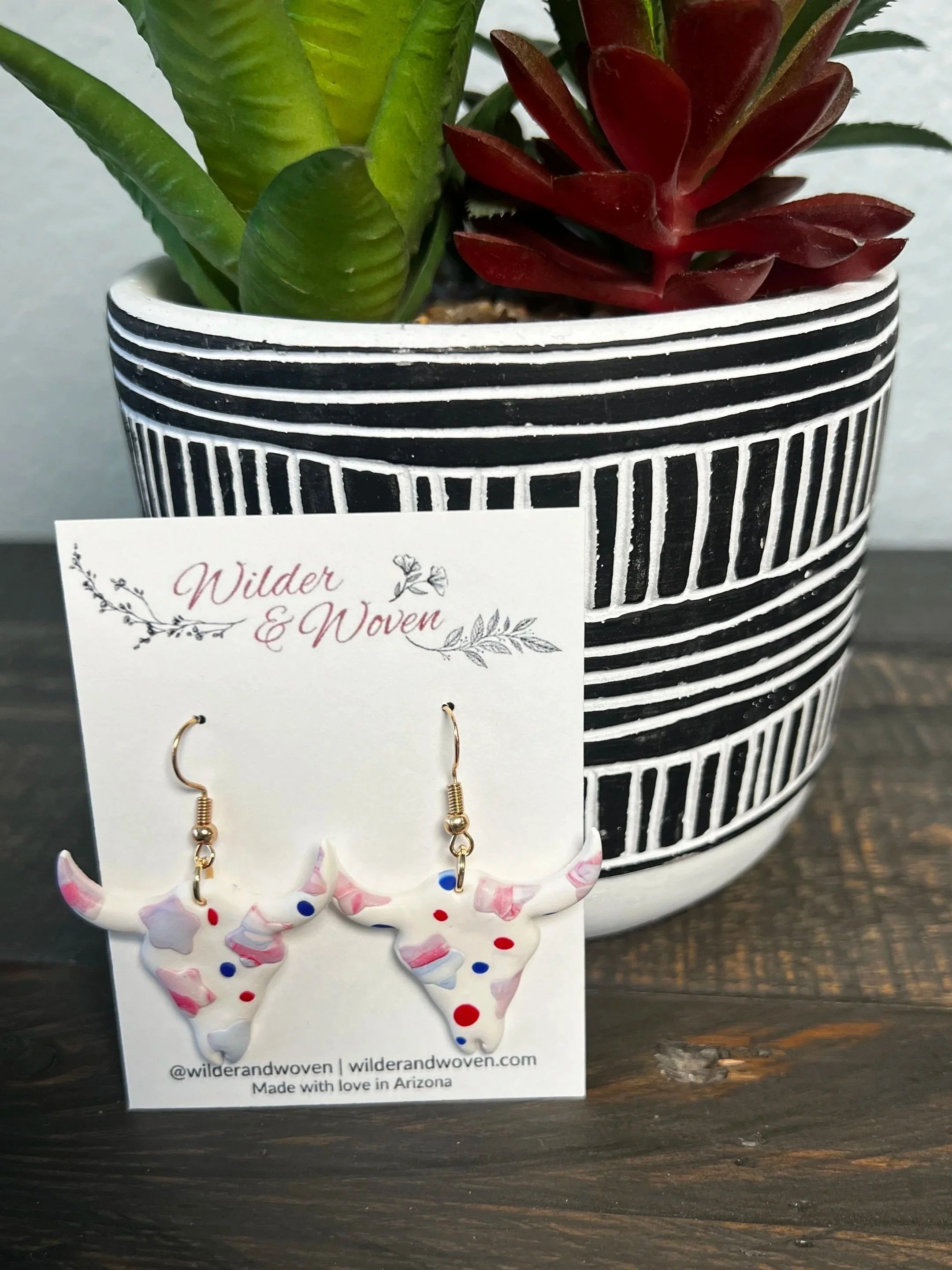 Handmade white polymer clay longhorn earrings with marbled red, white, and blue accents hanging from gold hooks on a Wilder & Woven display card.